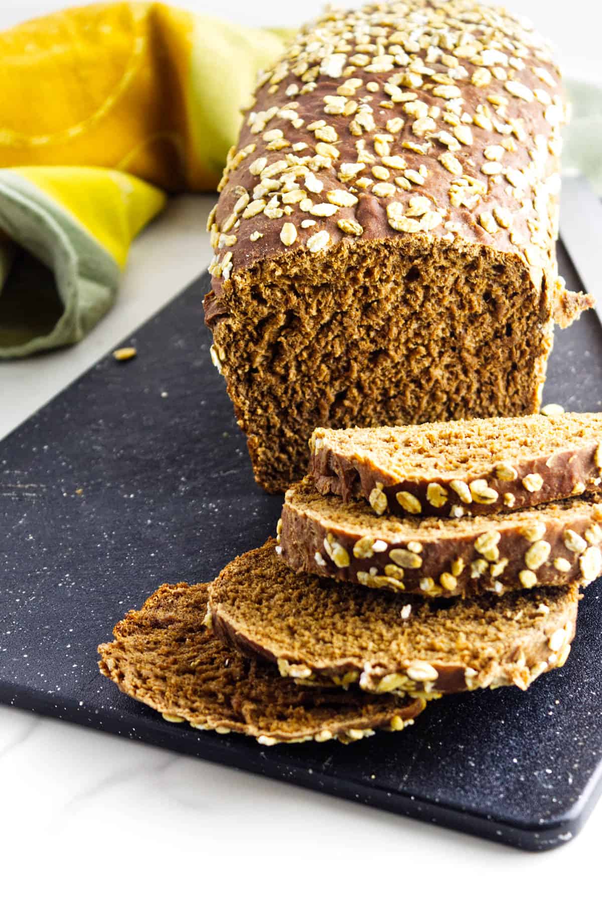 A loaf of sweet pumpernickle bread topped with oats sits on a black cutting board, several slices cut in front. A green and yellow towel adds color to the background.