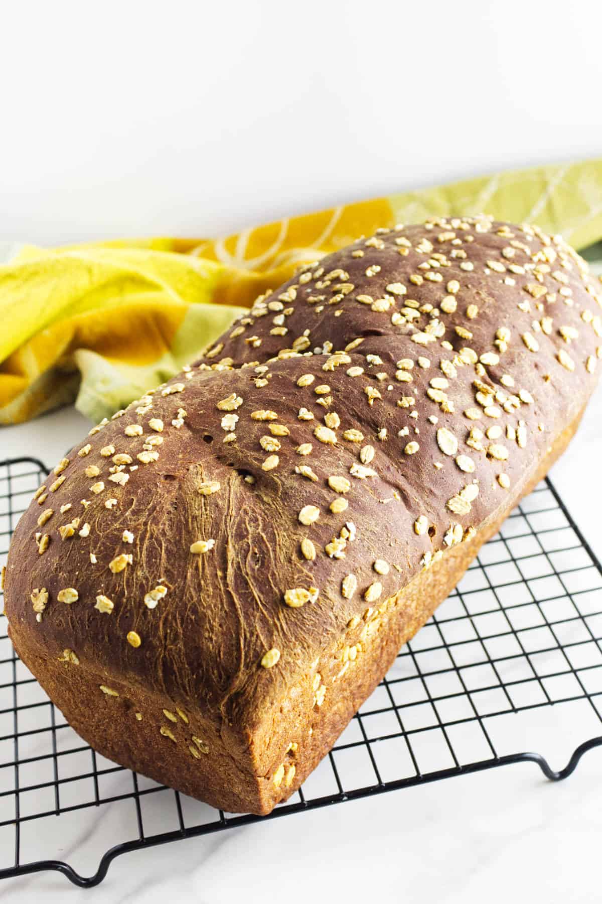 A loaf of oat-topped whole wheat bread, reminiscent of Cheesecake Factory brown bread, rests on a black cooling rack, with a yellow and green kitchen towel in the background.