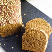 A loaf of homemade Cheesecake Factory brown bread topped with oats sits on a black cutting board. Two slices have been cut and are lying next to the loaf, with a green and yellow cloth partially visible in the background.