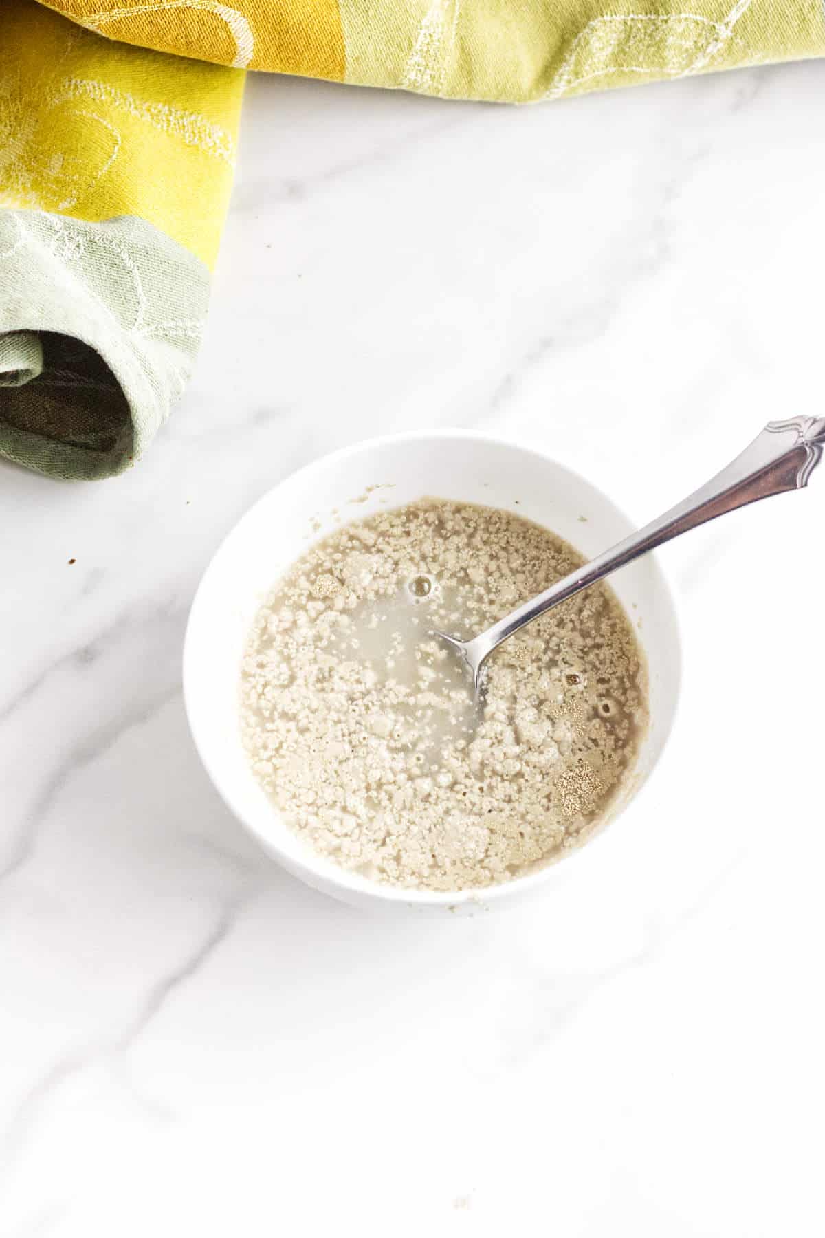 A white bowl with activated yeast mixture and a spoon inside, sitting on a white marble surface-perfect for making soft pumpernickle rolls or even those famous Cheesecake Factory brown dinner rolls. A yellow and green kitchen towel is partially visible in the background.