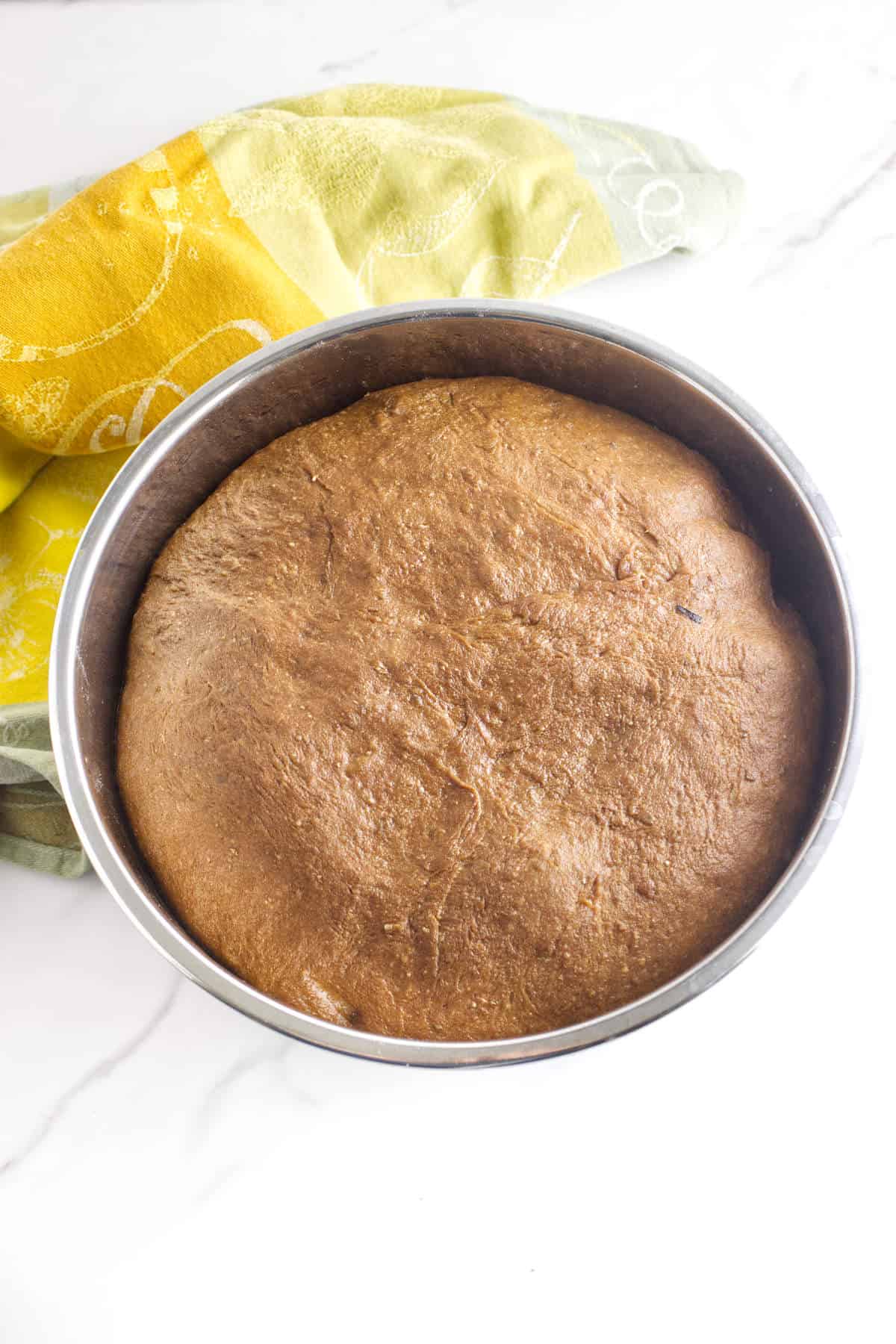 A round metal bowl filled with risen brown dough, reminiscent of Cheesecake Factory brown dinner rolls, sits on a white surface, partially covered by a green and yellow patterned cloth.