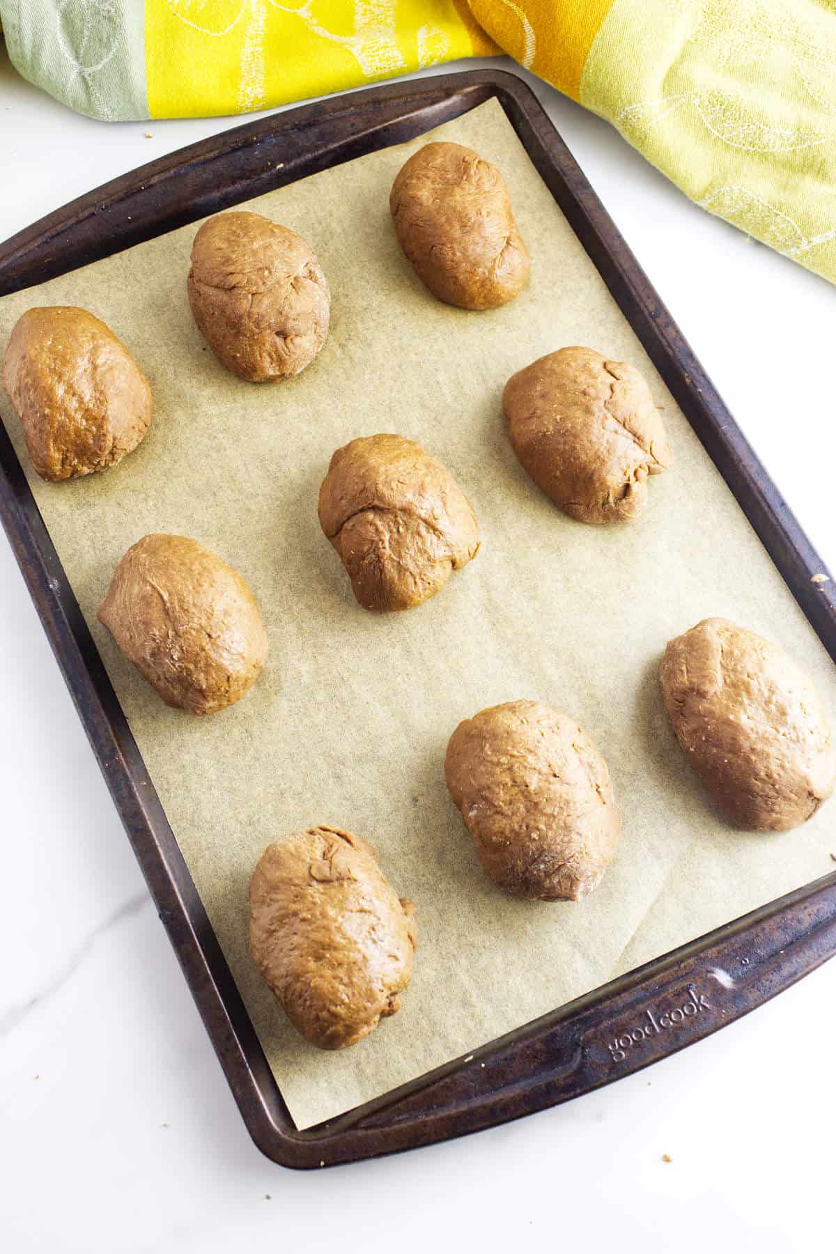 A baking sheet lined with parchment paper holds nine unbaked pumpernickle rolls spaced evenly apart. A yellow and green kitchen towel is partially visible in the background.