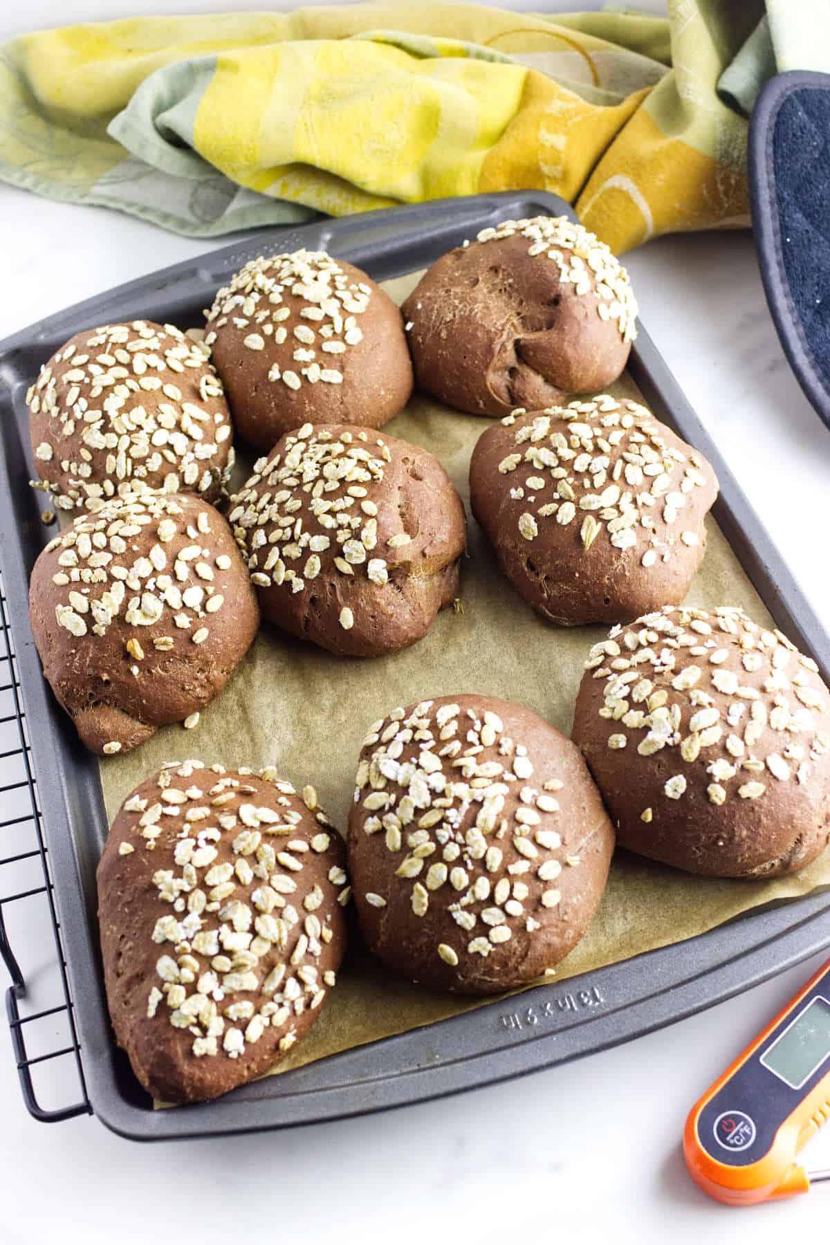 Eight round, dark brown pumpernickle rolls topped with oats sit on a parchment-lined baking sheet. A colorful towel and oven mitt are nearby, with a cooking thermometer at the bottom right corner of the image.