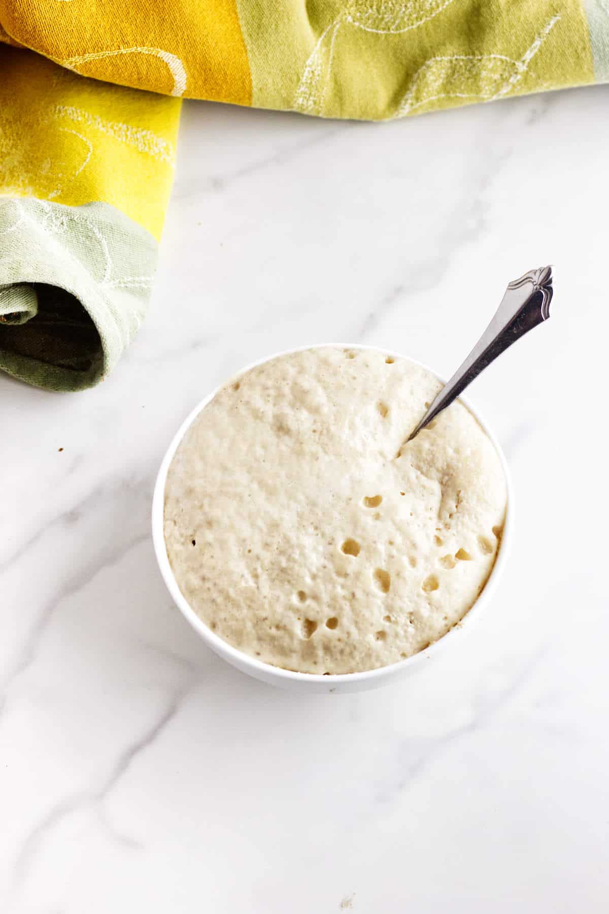 A white bowl filled with bubbly, risen bread dough-reminiscent of Cheesecake Factory brown dinner rolls-sits on a marble surface. A spoon is inserted into the dough, and a yellow and green cloth is partially visible in the background.