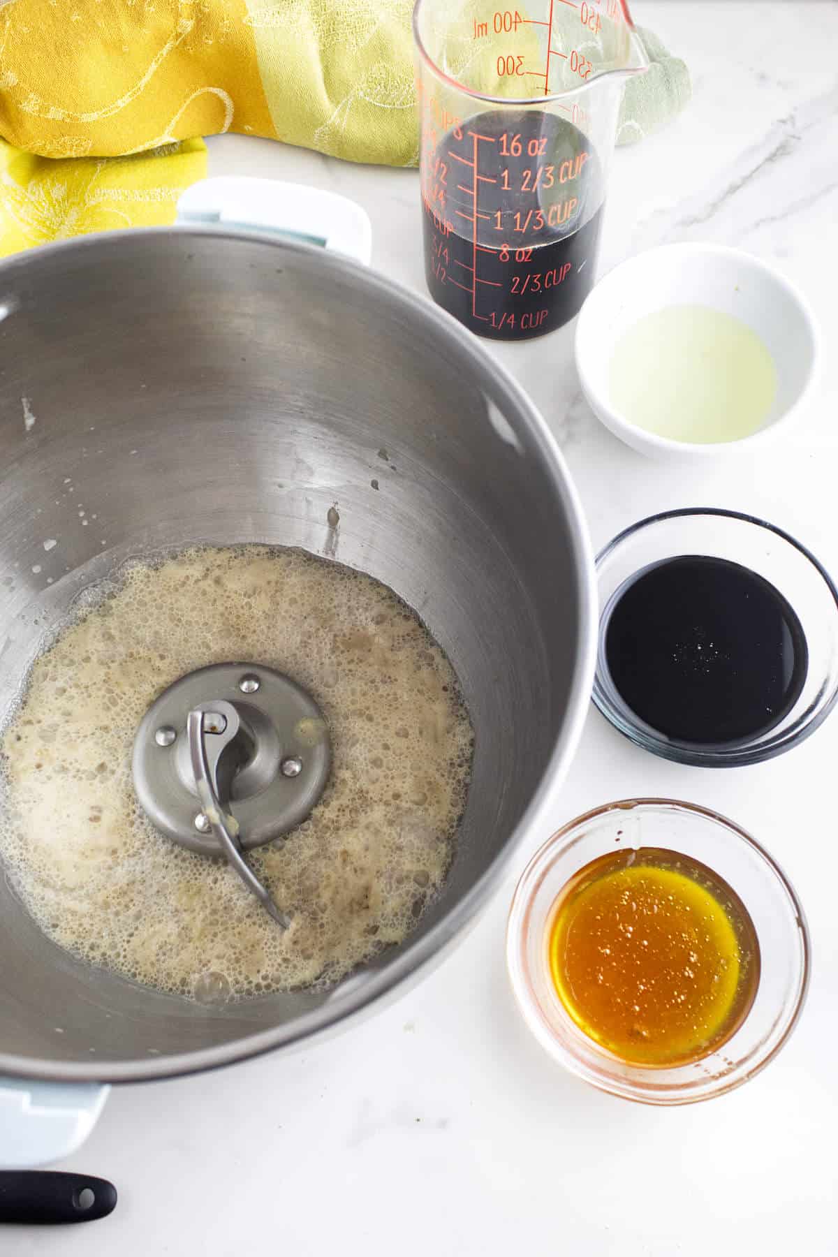 A mixing bowl with foamy liquid sits next to small bowls containing oil, honey, and molasses—classic ingredients for Cheesecake Factory brown dinner rolls or pumpernickle rolls—on a white countertop. A yellow cloth is in the background.