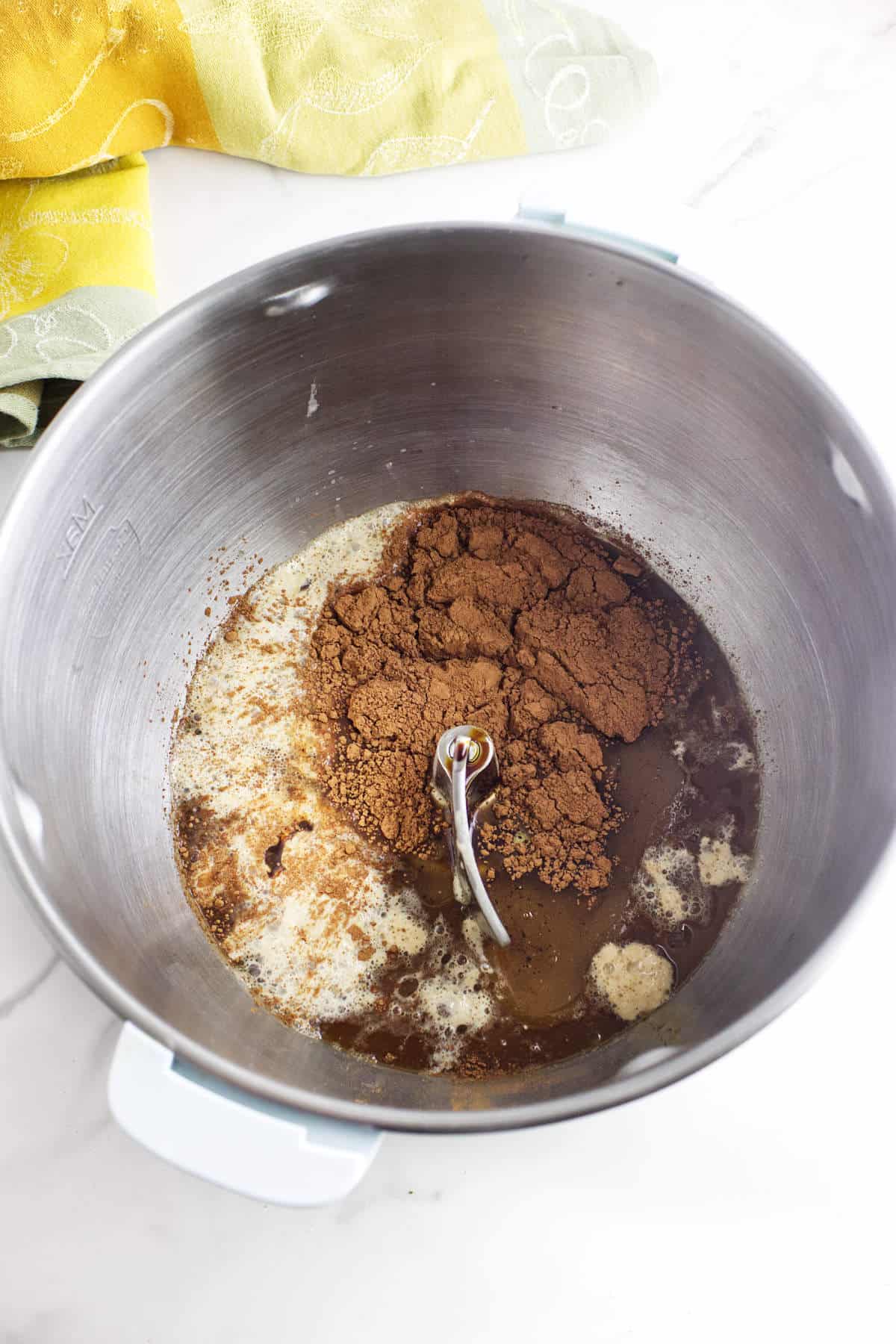 A mixing bowl with a stand mixer attachment holds cocoa powder, melted butter, sugar, and liquid ingredients-perfect for creating rich treats like Cheesecake Factory brown dinner rolls. A yellow and green kitchen towel sits on the white countertop.