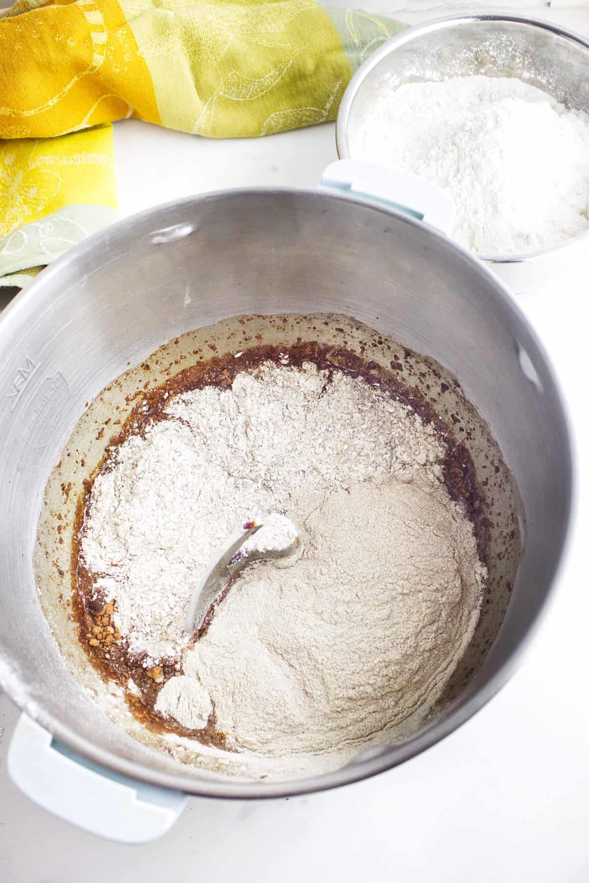 A mixing bowl with flour and wet ingredients partially combined for homemade Cheesecake Factory brown dinner rolls, with a spoon inside. In the background, a bowl of flour and a yellow and green kitchen towel rest on a white countertop.