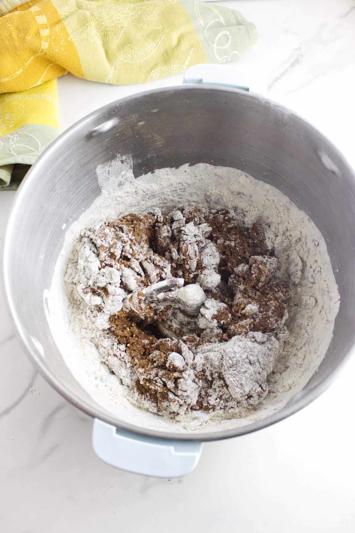 A metal mixing bowl containing partially mixed cookie dough and flour sits on a white countertop, with a metal spoon inside and a yellow and green kitchen towel nearby, reminiscent of making Cheesecake Factory brown dinner rolls at home.