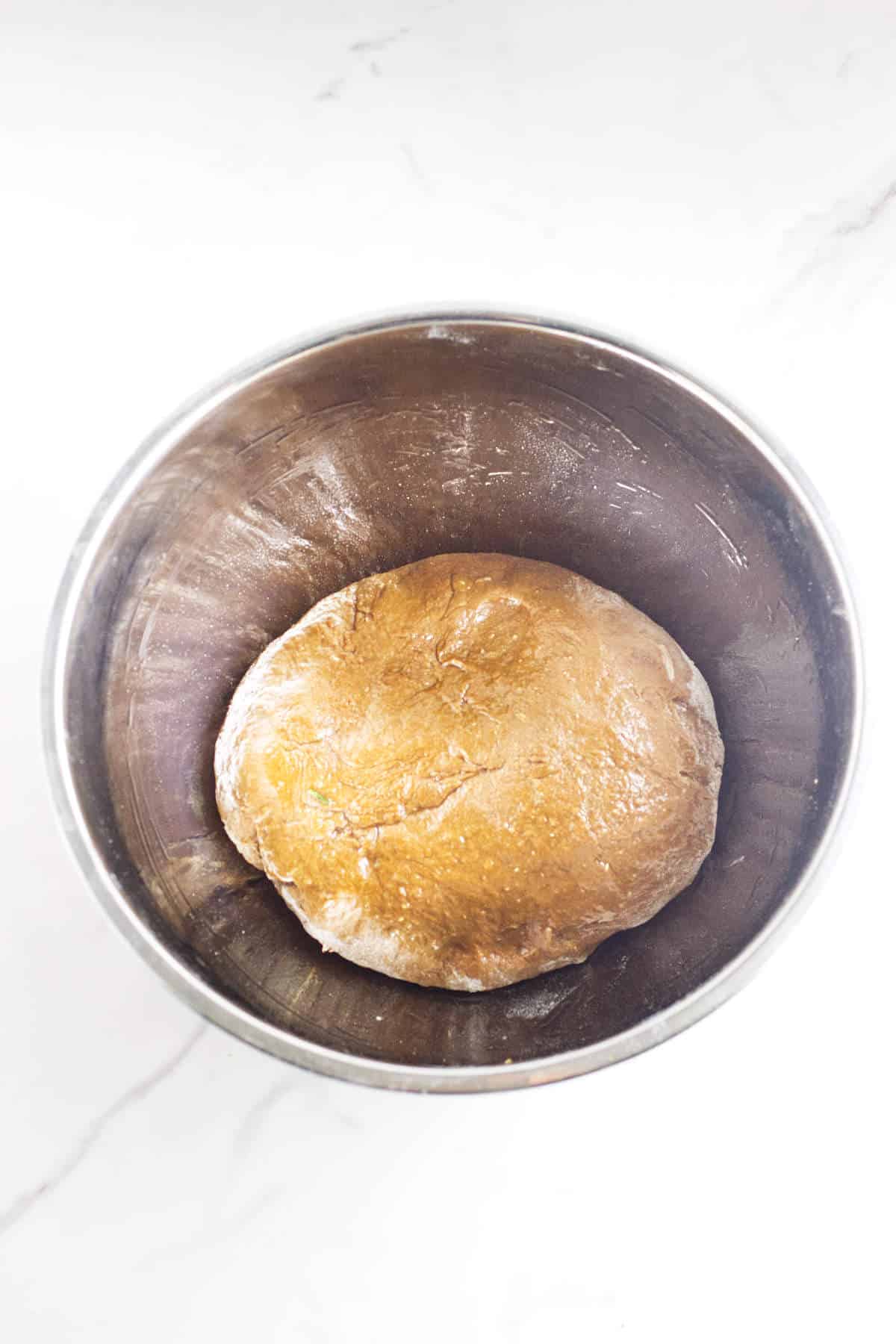 A ball of dough, ready to become delicious pumpernickle rolls, rests in a stainless steel mixing bowl on a white marble surface.