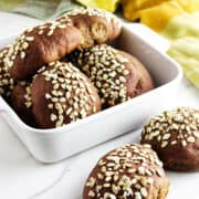 A white dish filled with several round, dark brown pumpernickle rolls topped with oats, with two rolls placed beside the dish on a white surface. A yellow and green cloth is in the background, reminiscent of Cheesecake Factory brown dinner rolls.