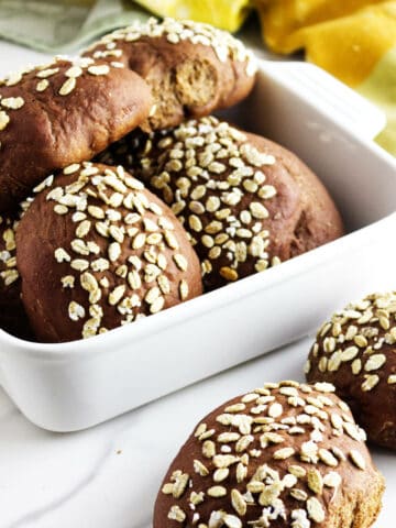 A white dish filled with several round, dark brown pumpernickle rolls topped with oats, with two rolls placed beside the dish on a white surface. A yellow and green cloth is in the background, reminiscent of Cheesecake Factory brown dinner rolls.