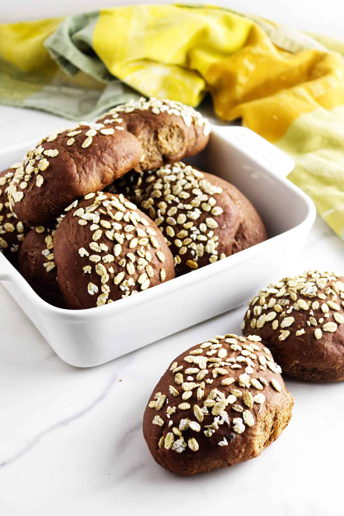 A white dish filled with dark brown pumpernickle rolls topped with oats, reminiscent of Cheesecake Factory brown dinner rolls, with two additional rolls in front. A yellow and green towel is in the background on a white surface.