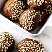 A white dish filled with round, dark brown pumpernickle rolls topped with oat flakes sits on a white marble surface. Two rolls, reminiscent of Cheesecake Factory brown dinner rolls, are placed outside the dish.