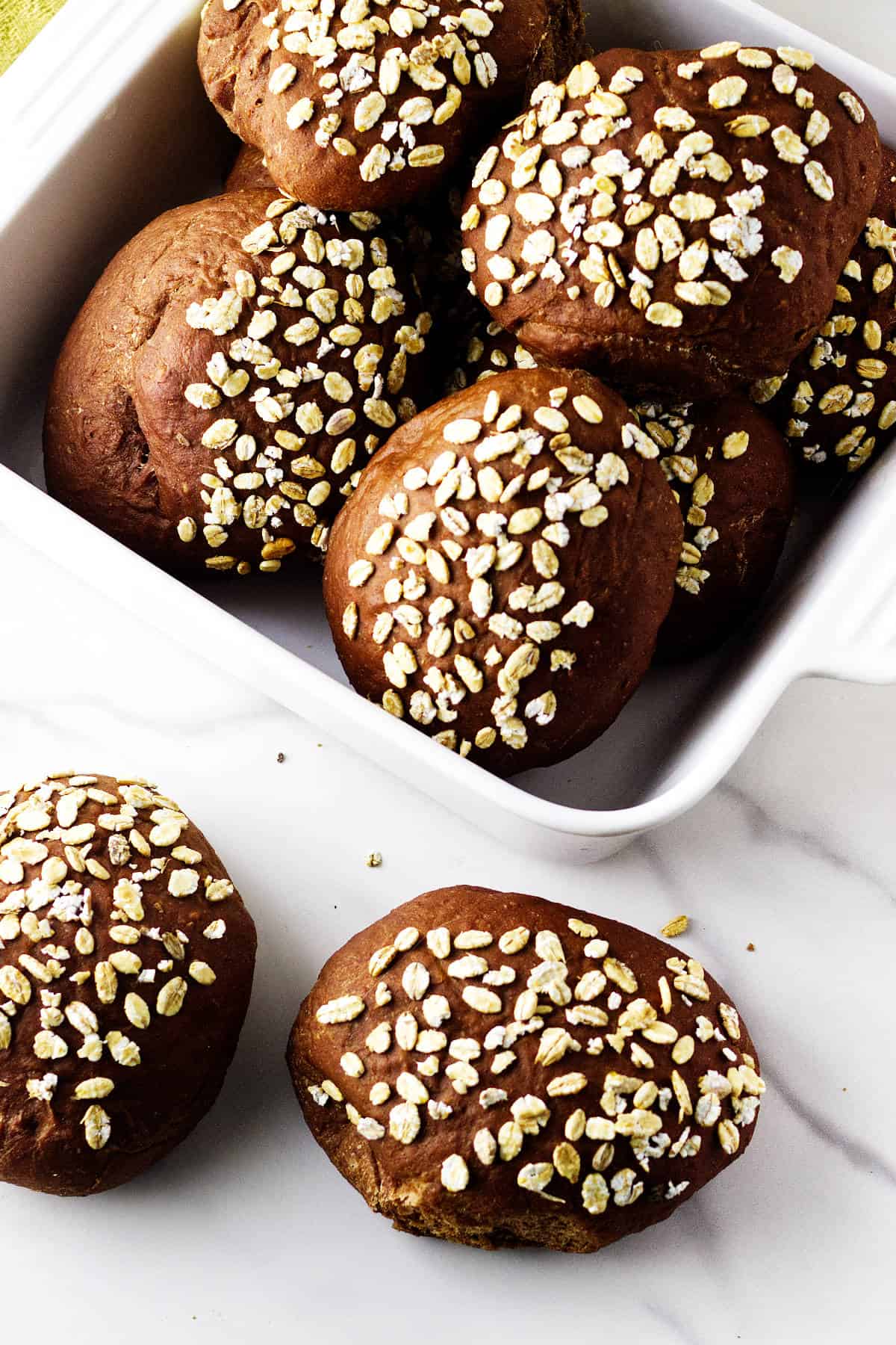 A white dish filled with round, dark brown pumpernickle rolls topped with oat flakes sits on a white marble surface. Two rolls, reminiscent of Cheesecake Factory brown dinner rolls, are placed outside the dish.