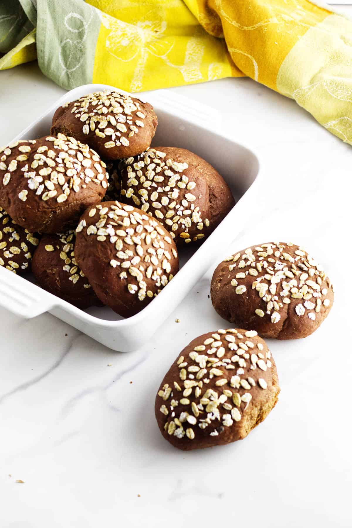 A white baking dish filled with round, Cheesecake Factory brown dinner rolls topped with oats sits on a white surface. Two rolls rest outside the dish, while a yellow and green kitchen towel adds color in the background.