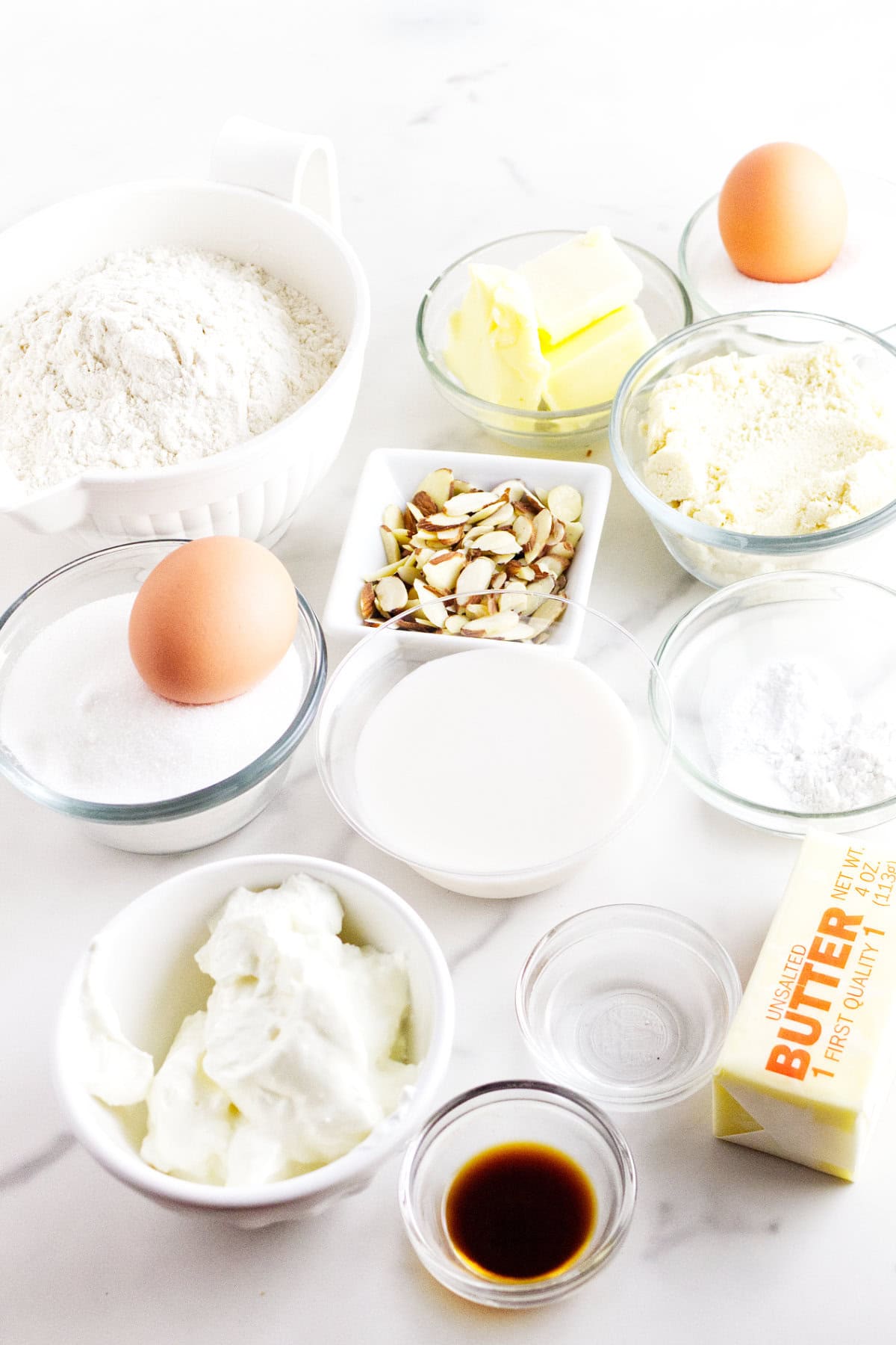 Assorted baking ingredients on a white surface, perfect for making almond muffins or frangipane muffins, including flour, sugar, almonds, butter, eggs, milk, sour cream, vanilla extract, and baking powder in various bowls and containers.