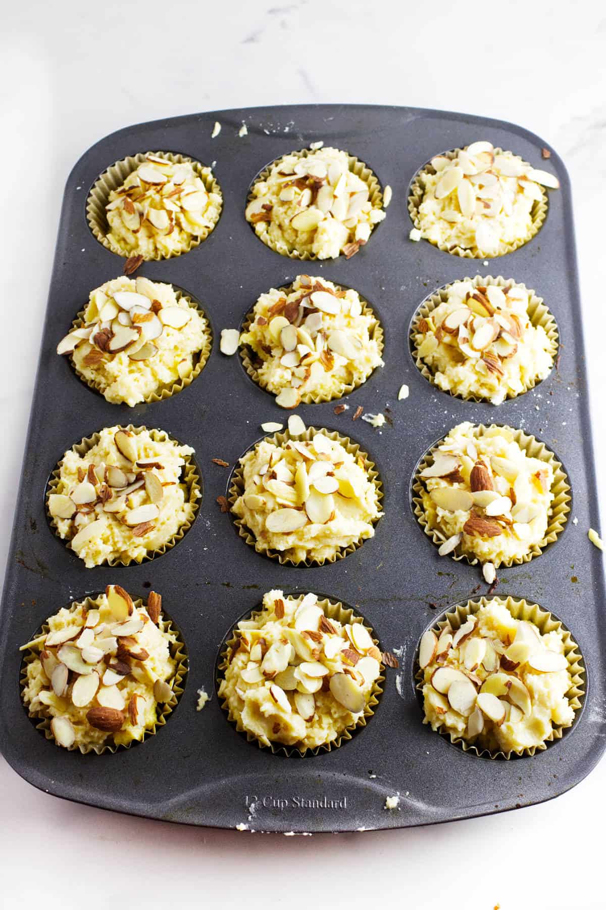 A muffin tin filled with almond muffins topped with sliced almonds sits on a white surface. Prepared in paper liners, these almond-filled muffins are ready to be baked.