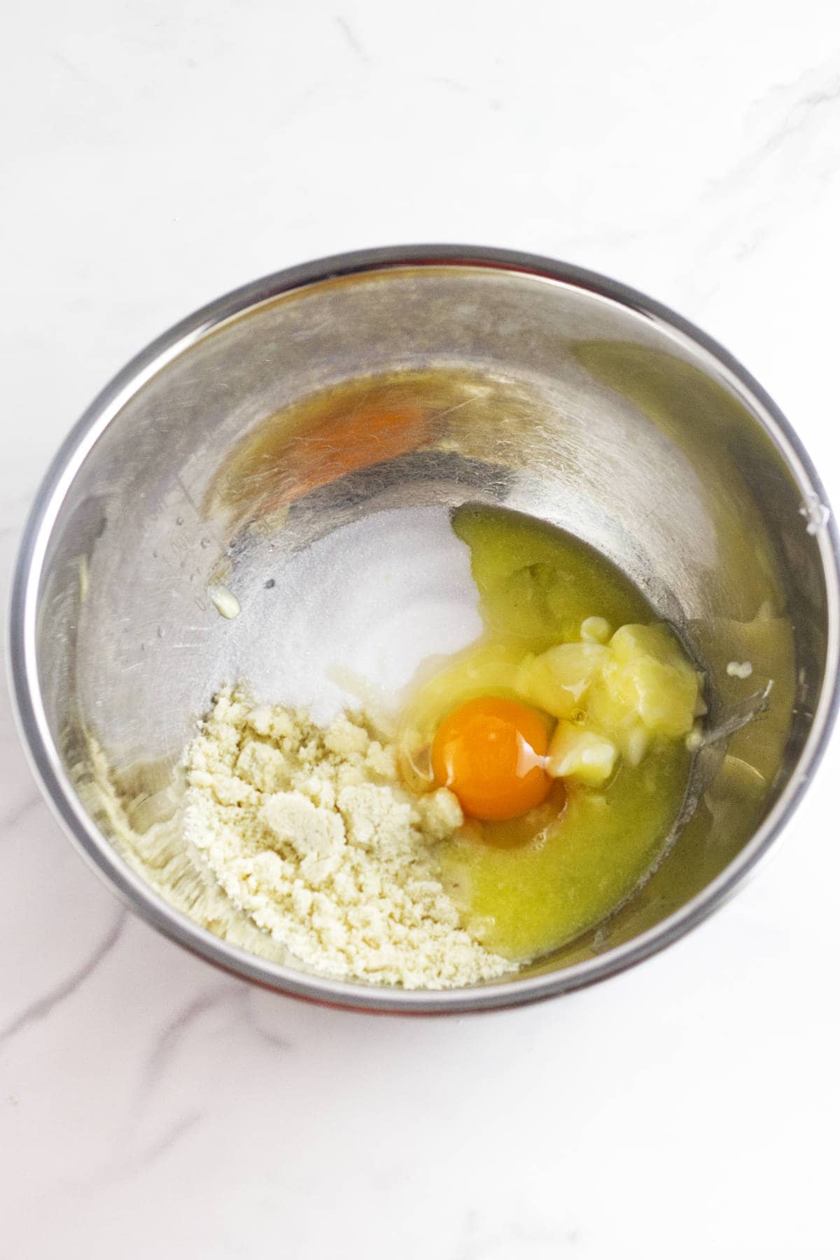 A metal mixing bowl containing almond flour, an egg, softened butter, and sugar on a white marble surface—perfect for making delicious frangipane muffins.