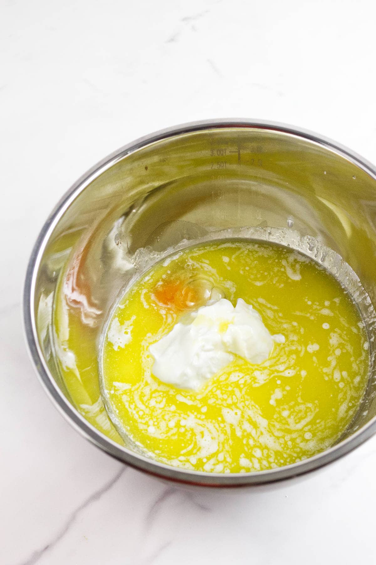A metal mixing bowl containing melted butter, a cracked egg, and a dollop of yogurt on a white marble surface-perfect beginnings for homemade frangipane muffins.