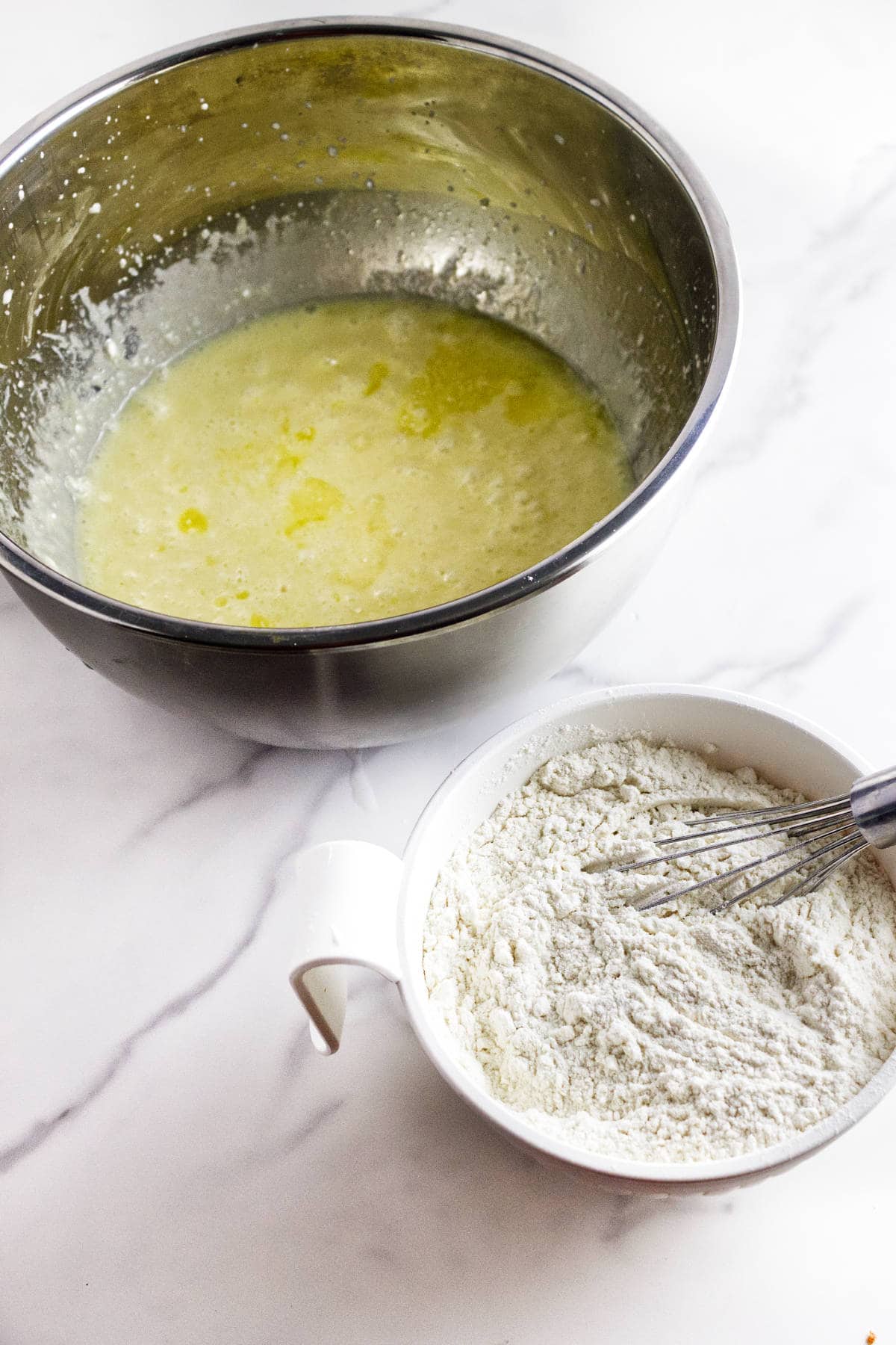 A metal bowl with wet batter for almond muffins being whisked, alongside a white bowl of flour, both resting on a white marble countertop.
