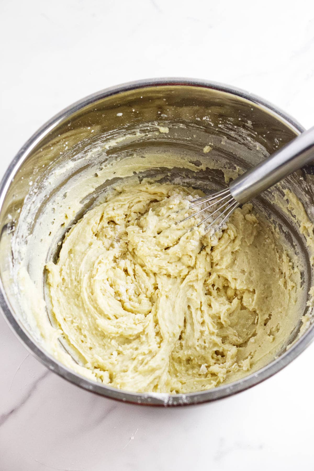 A metal mixing bowl filled with thick, pale batter for almond muffins is being mixed with a wire whisk, sitting on a white marble surface.