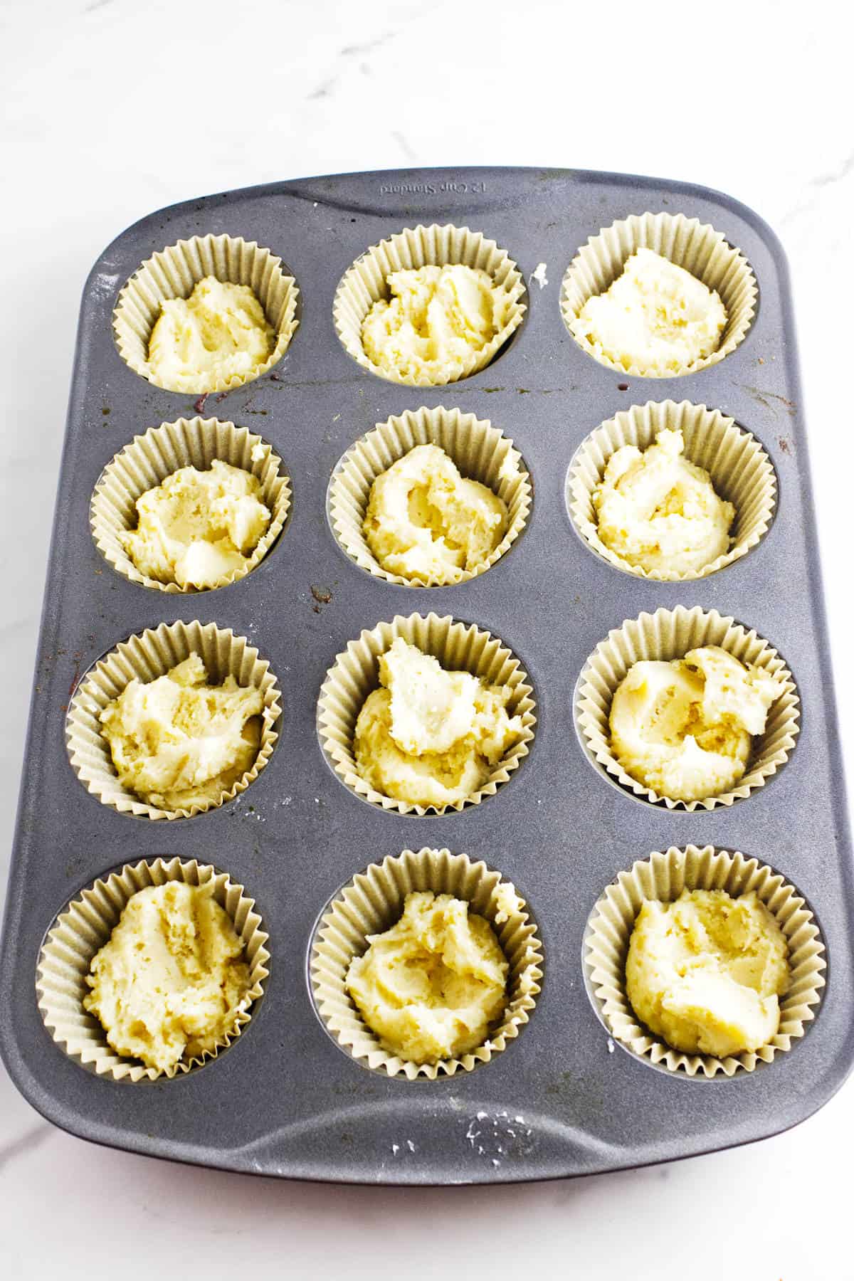 A muffin tin with twelve paper liners, each filled with unbaked almond muffins batter, sits on a white surface. The batter appears plain with a slightly rough texture.
