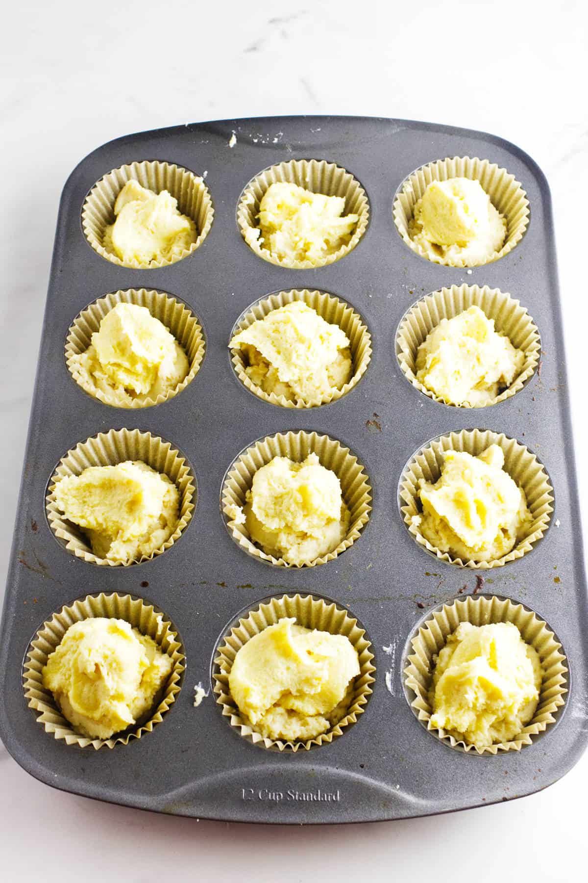 A muffin tin lined with paper cups, each filled with scoops of raw almond muffin batter, sits on a white countertop ready for baking.