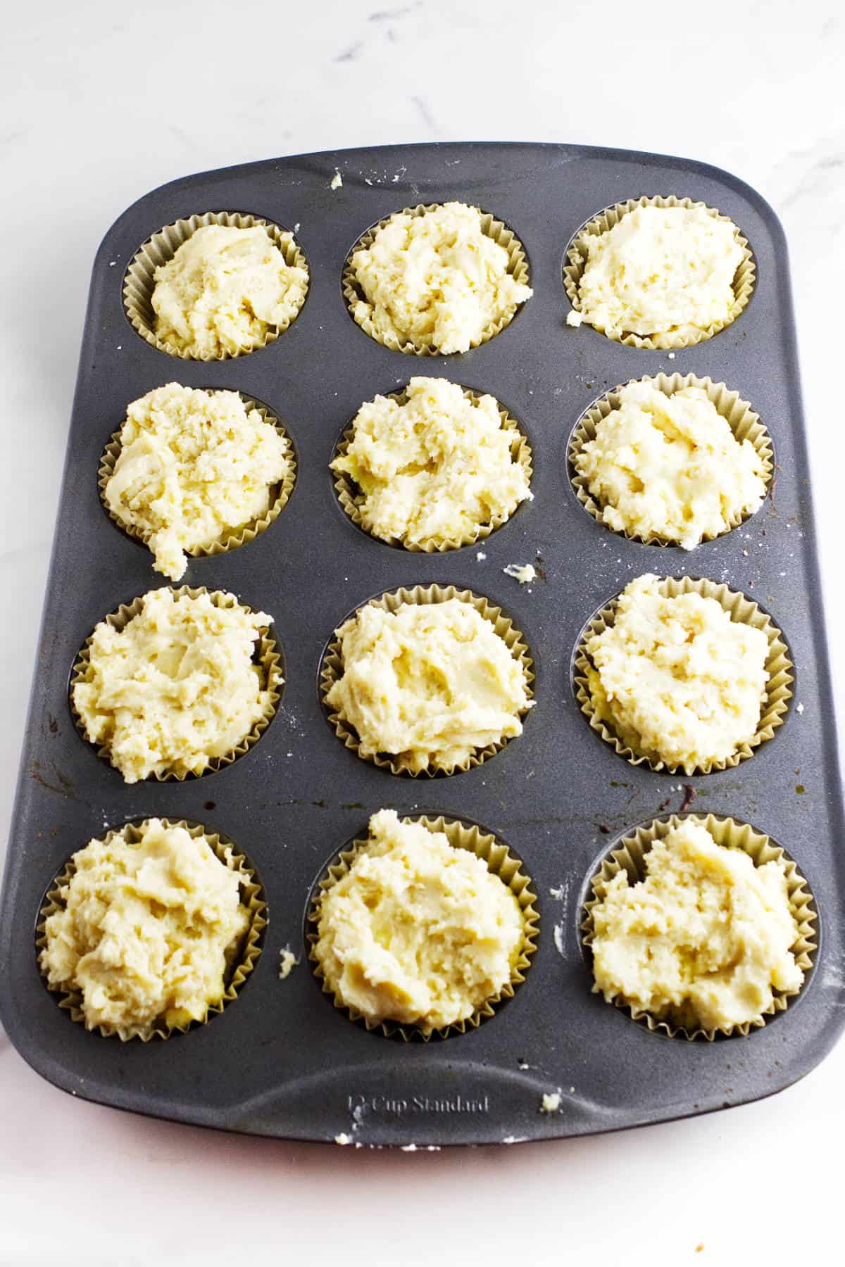 A 12-cup muffin tin filled with raw, scooped almond muffin batter in paper liners, ready to be baked, sits on a white surface.
