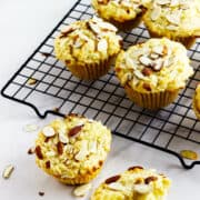 Six almond muffins, some almond filled, rest on a black cooling rack, with a few scattered almond slices on the white surface nearby. The frangipane muffins are golden brown and appear light and fluffy.