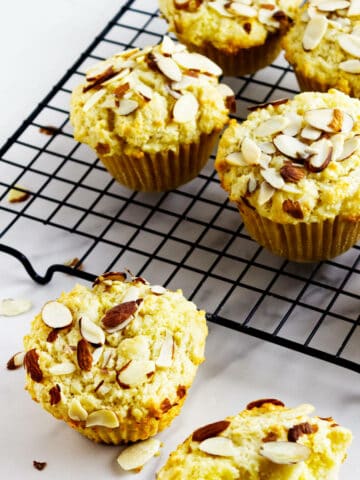 Six almond muffins, some almond filled, rest on a black cooling rack, with a few scattered almond slices on the white surface nearby. The frangipane muffins are golden brown and appear light and fluffy.