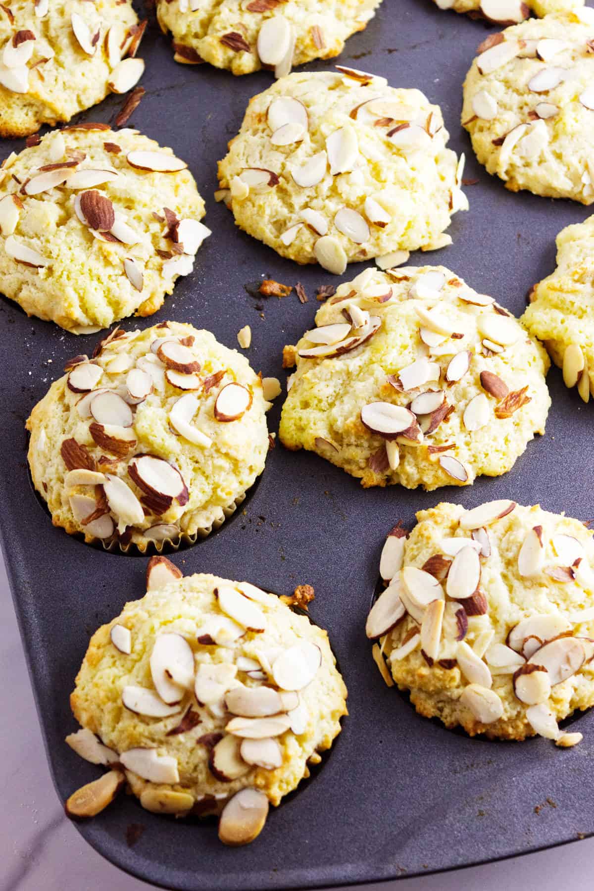 A muffin tin filled with freshly baked almond muffins topped with sliced almonds, some slightly golden, arranged closely together.