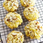 Six almond muffins, some almond-filled, topped with sliced almonds are cooling on a black wire rack, with a few almond slices scattered around on a white surface.