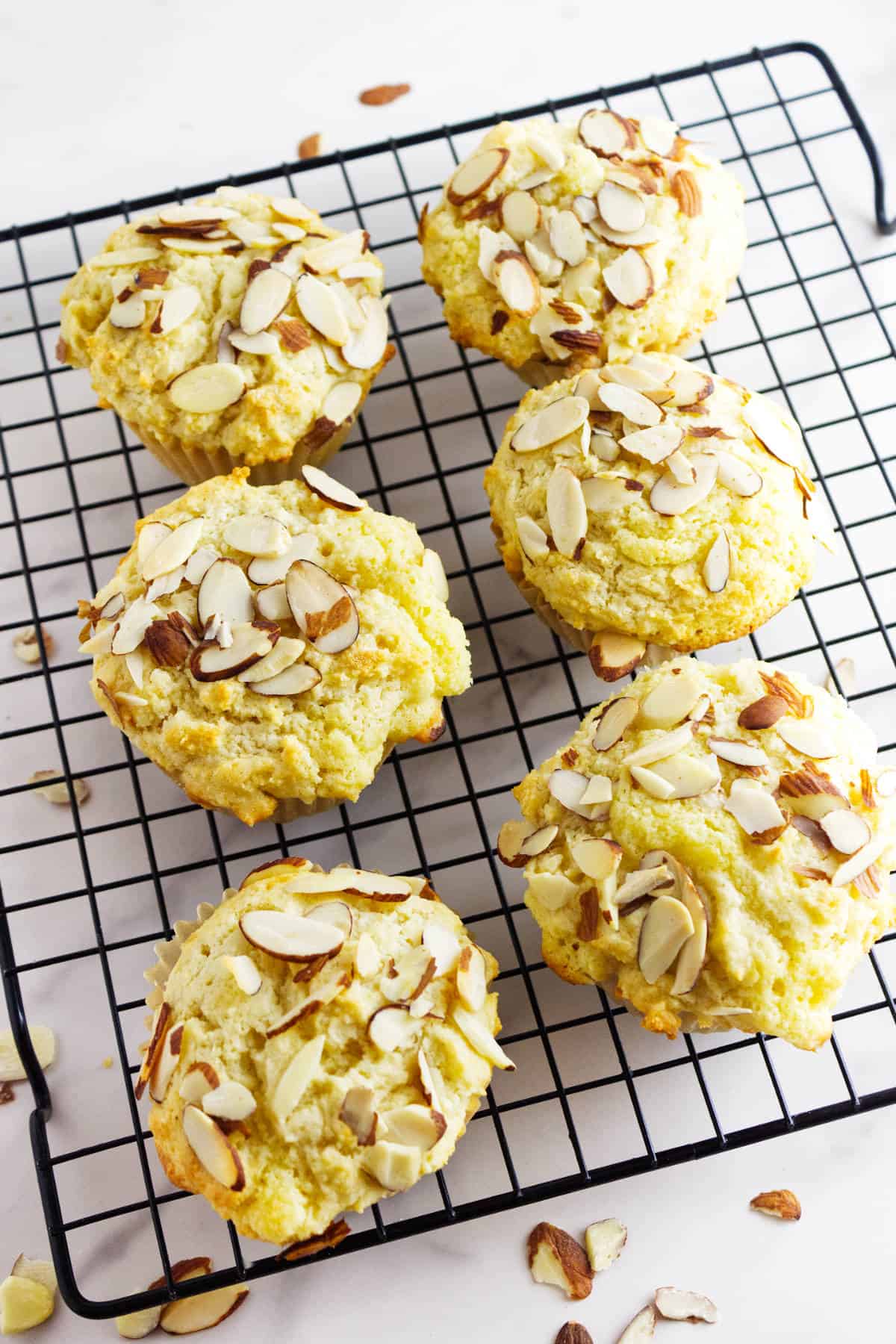 Six almond muffins, some almond-filled, topped with sliced almonds are cooling on a black wire rack, with a few almond slices scattered around on a white surface.