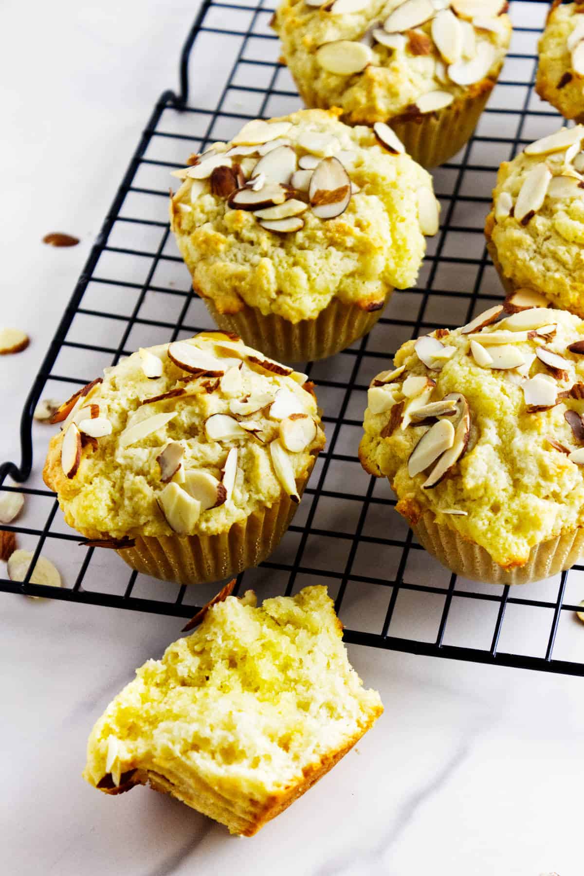 Almond muffins, like these tempting frangipane muffins topped with sliced almonds, are cooling on a black wire rack. One almond-filled muffin reveals its soft, fluffy interior, while scattered almond slices decorate the white surface.