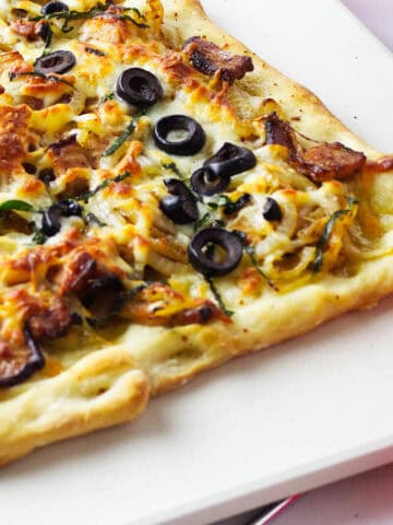 A close-up of a homemade flatbread pizza with melted cheese, sliced black olives, mushrooms, and herbs on a golden, crispy crust, served on a white surface.