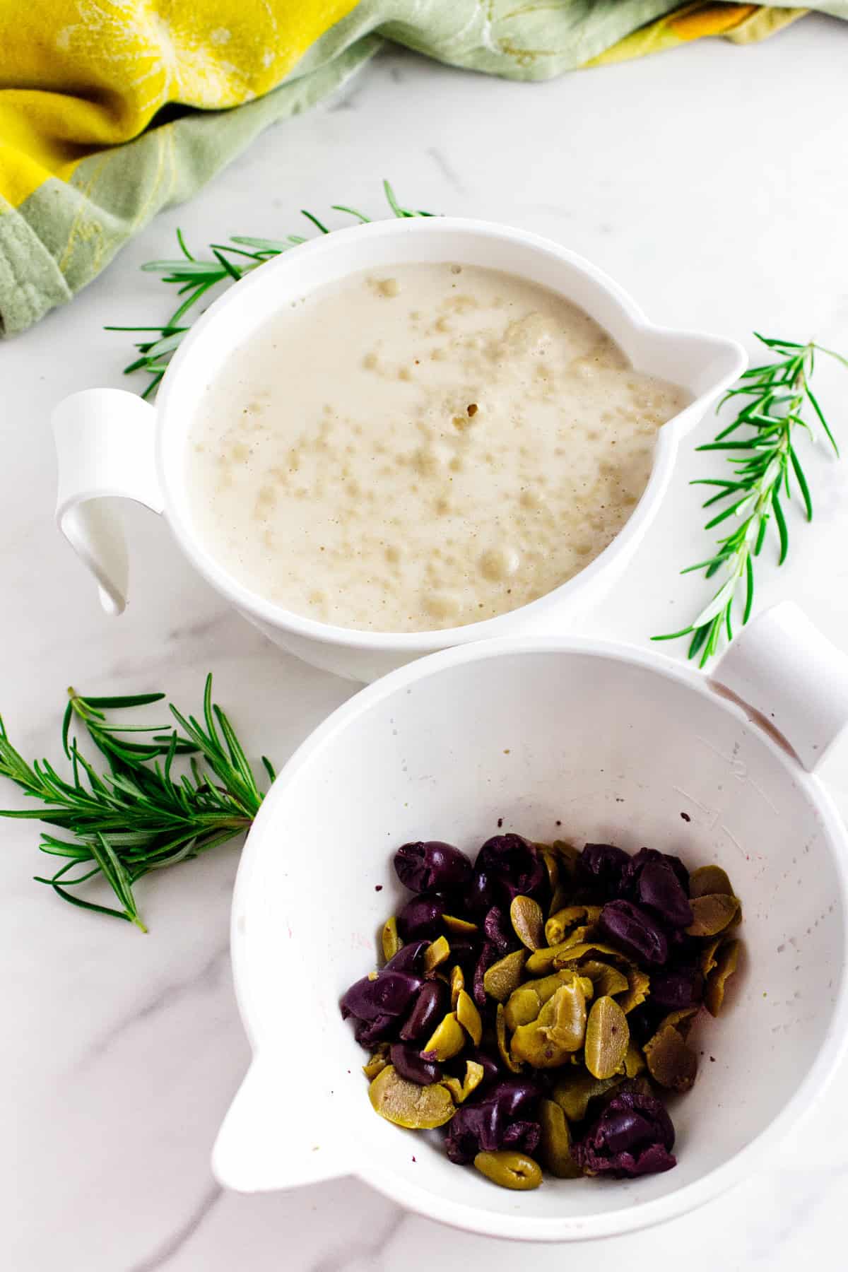 Two white bowls on a marble surface-one with a creamy, beige mixture perfect for pairing with no knead baguettes, the other filled with chopped green and black olives. Fresh rosemary sprigs and a yellow-green cloth complete the scene.