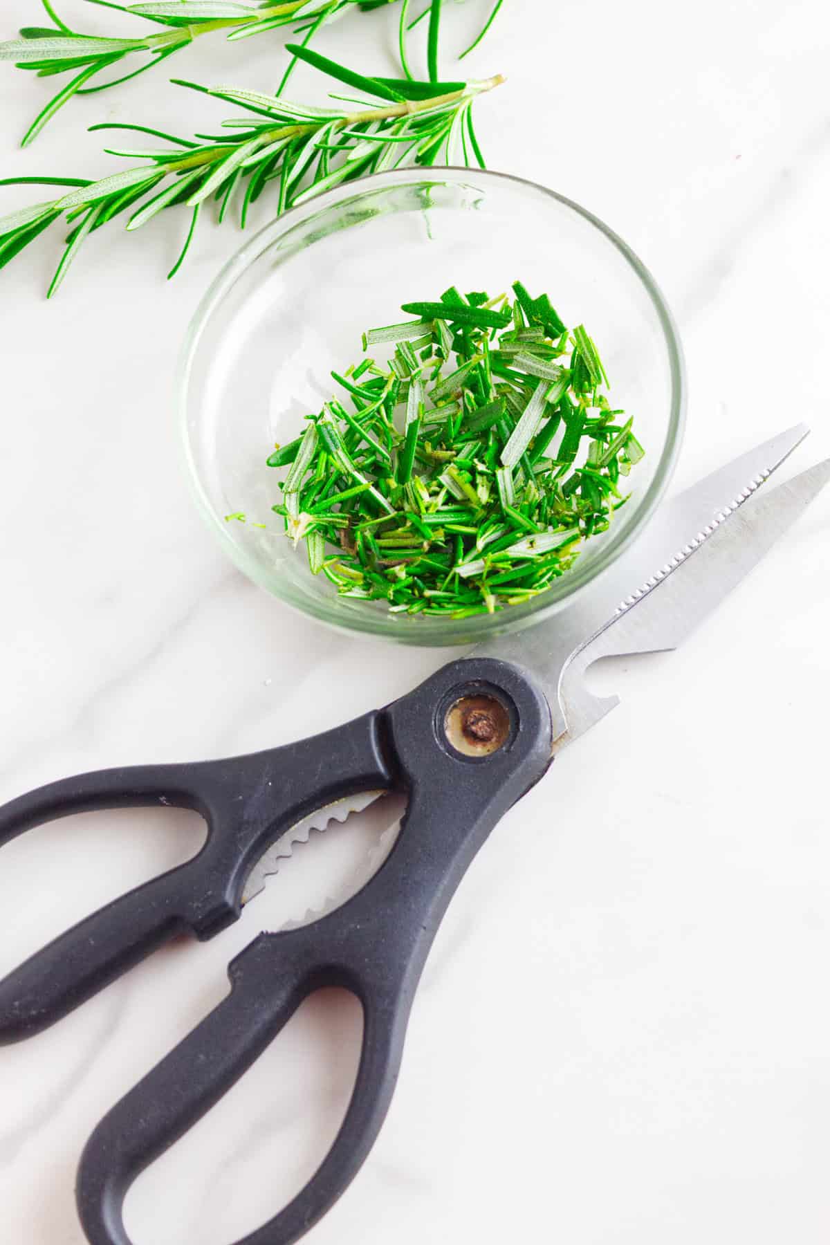 A pair of black kitchen scissors lies next to a small glass bowl filled with freshly chopped rosemary on a white surface, perfect for flavoring no knead baguettes, with sprigs of rosemary nearby.