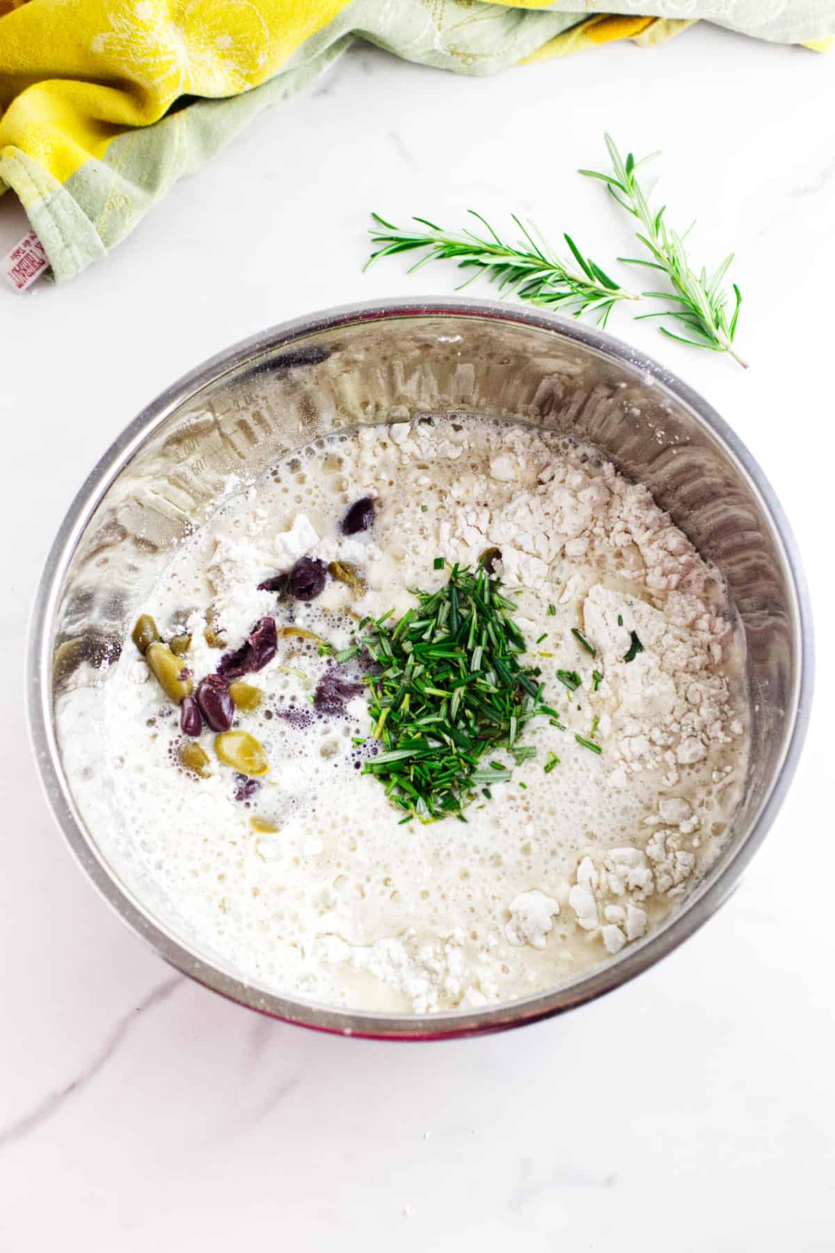 A mixing bowl for no knead baguettes sits on a white countertop, filled with flour, milk, chopped green herbs, olives, and feta cheese. Nearby are rosemary sprigs and a yellow cloth.