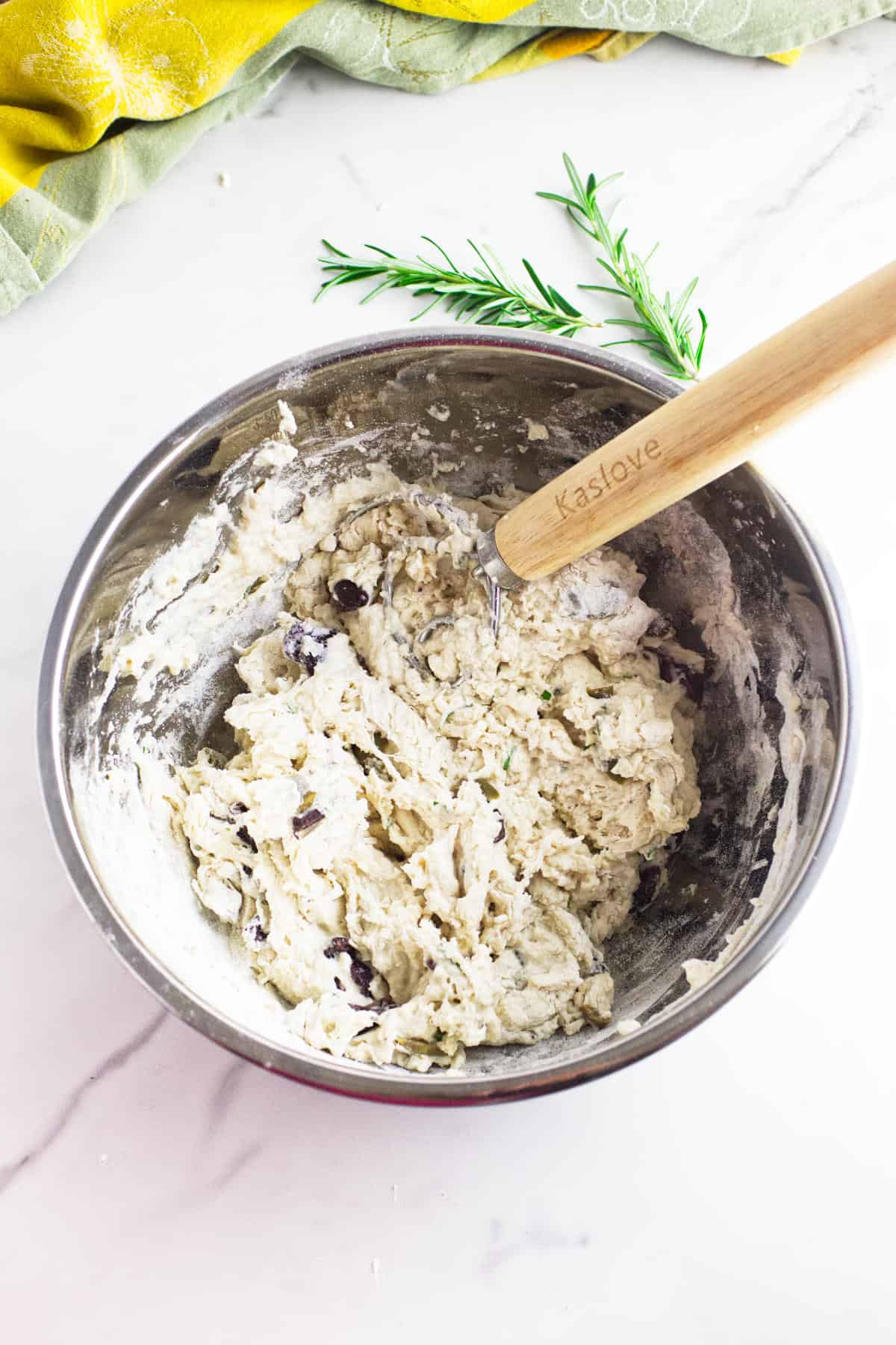 A metal mixing bowl filled with no knead baguette dough mixed with herbs and olives is being stirred with a wooden-handled pastry blender. A sprig of fresh rosemary and two cloths rest on the marble countertop beside the bowl.