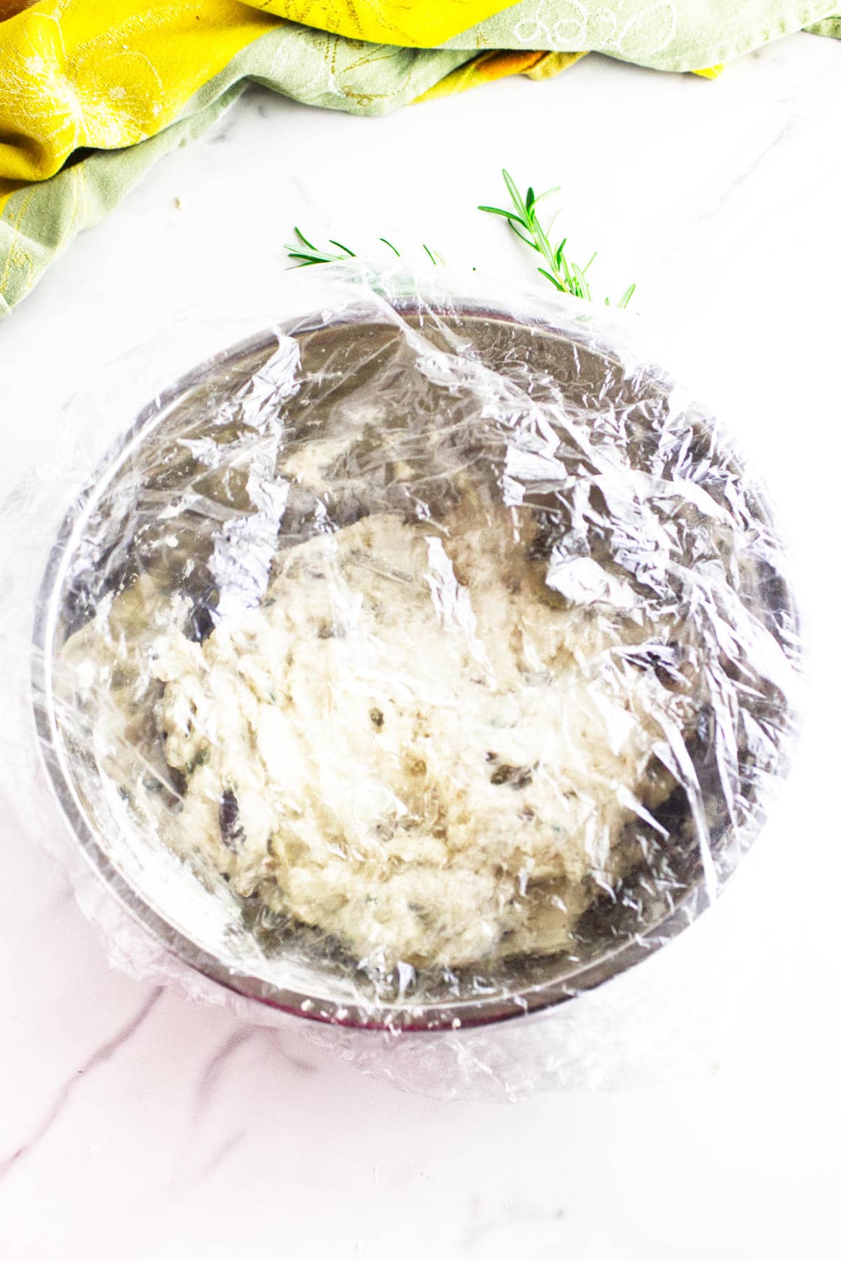A metal bowl of dough for no knead baguettes is covered with plastic wrap, resting on a white surface with sprigs of rosemary nearby and a yellow-green cloth at the top of the image.