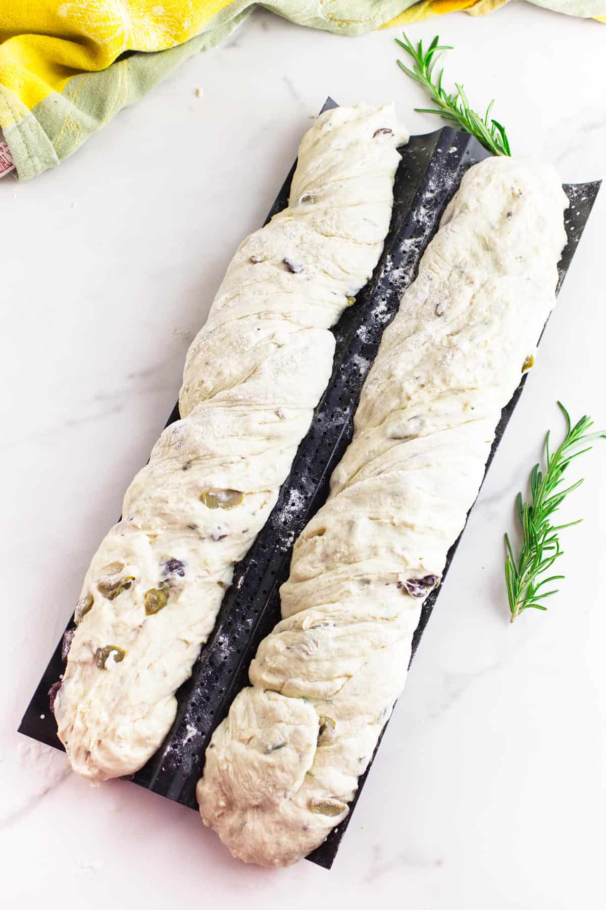 Two unbaked no knead baguette loaves with olives and herbs rest on a black baguette tray, topped with flour. A sprig of rosemary and a yellow-green cloth are nearby on a white marble surface.