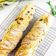 Two freshly baked no knead baguettes with visible olive pieces rest on a wire cooling rack. A sprig of rosemary and a green cloth are placed nearby on a white surface.