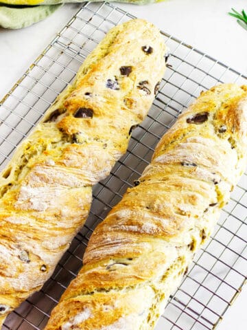 Two freshly baked no knead baguettes with visible olive pieces rest on a wire cooling rack. A sprig of rosemary and a green cloth are placed nearby on a white surface.