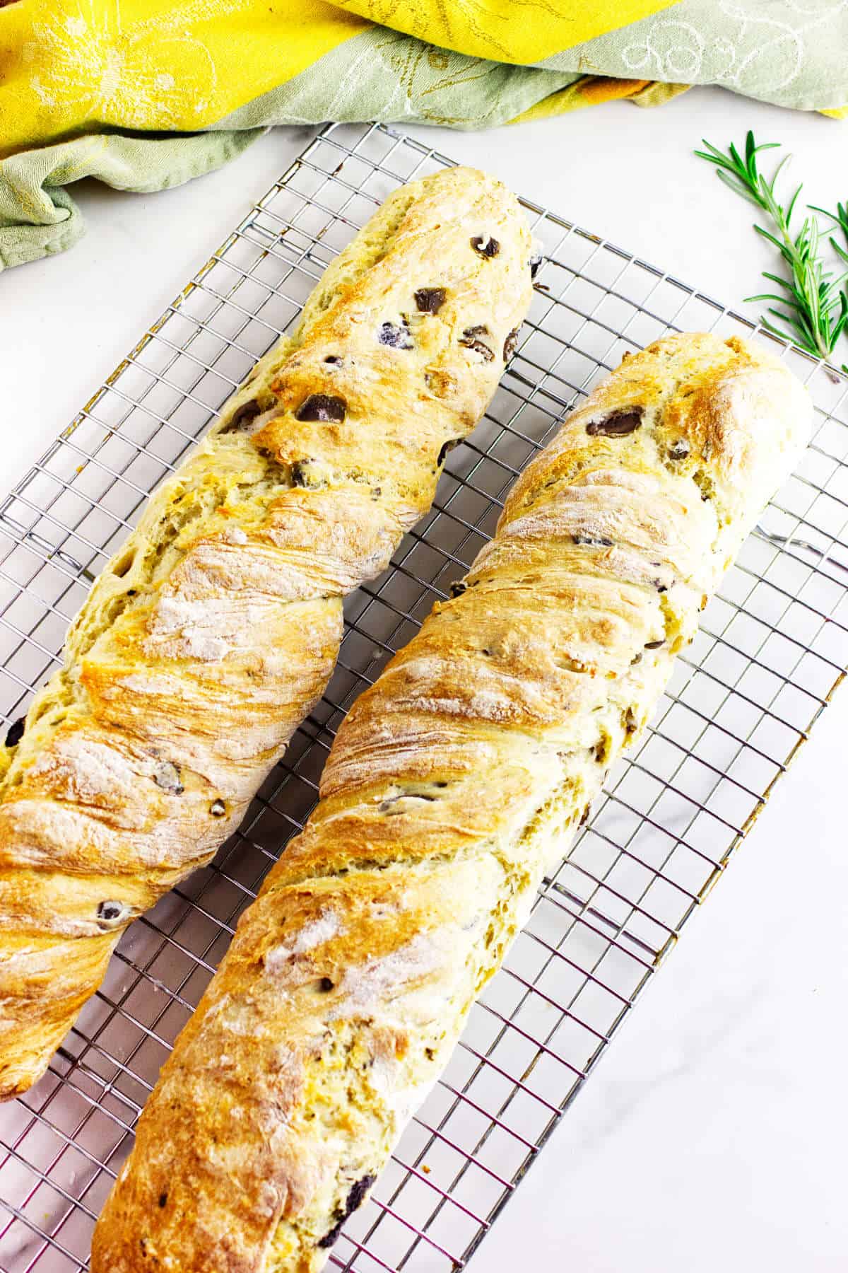 Two golden-brown loaves of homemade no knead baguettes cool on a wire rack, with a sprig of rosemary and yellow and green cloths in the background.