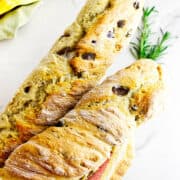 Two rustic loaves of no knead baguettes with olive and rosemary rest on a white surface, partially wrapped in a striped kitchen towel, with a sprig of rosemary and colorful cloths in the background.
