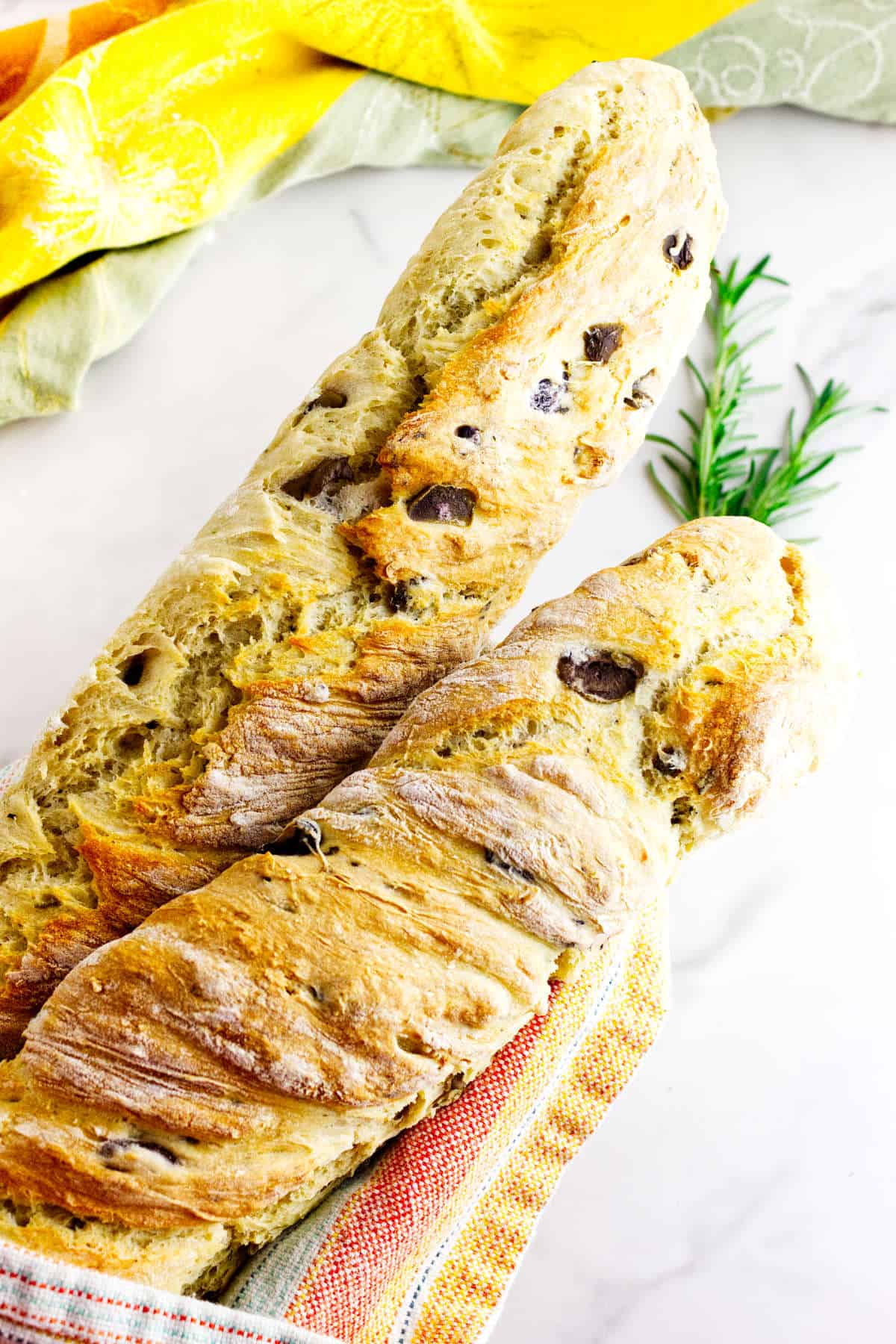 Two rustic loaves of no knead baguettes with olive and rosemary rest on a white surface, partially wrapped in a striped kitchen towel, with a sprig of rosemary and colorful cloths in the background.