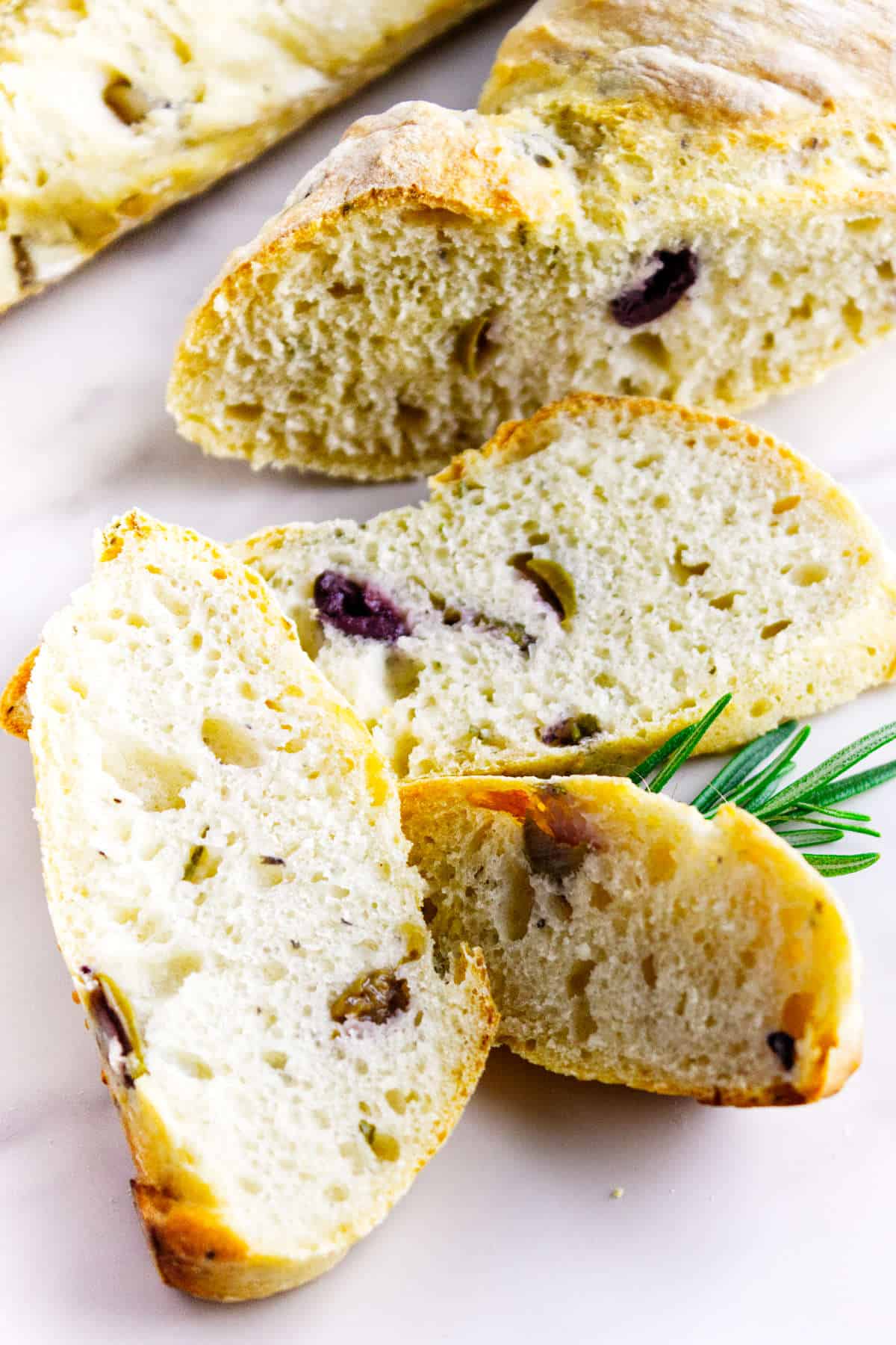 Sliced no knead baguettes with olives on a white surface, paired with a sprig of fresh rosemary. The bread features a light, airy texture and a golden crust.