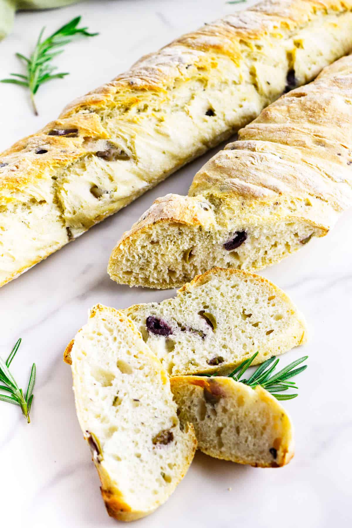 A sliced loaf of no knead baguettes with visible pieces of olives and herbs, displayed on a white surface with sprigs of rosemary.