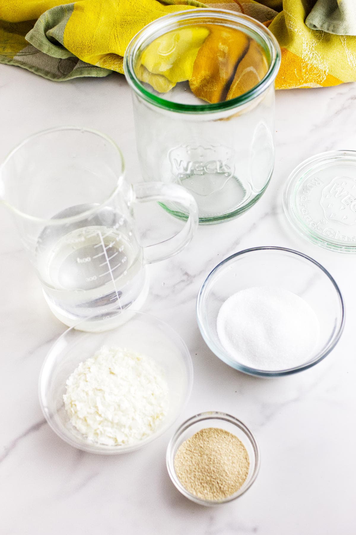 A glass jar, a measuring cup with water, a bowl of sugar, cornstarch, and dry yeast are arranged on a white countertop—perfect essentials for making a potato flake sourdough bread starter—with yellow and green cloths in the background.
