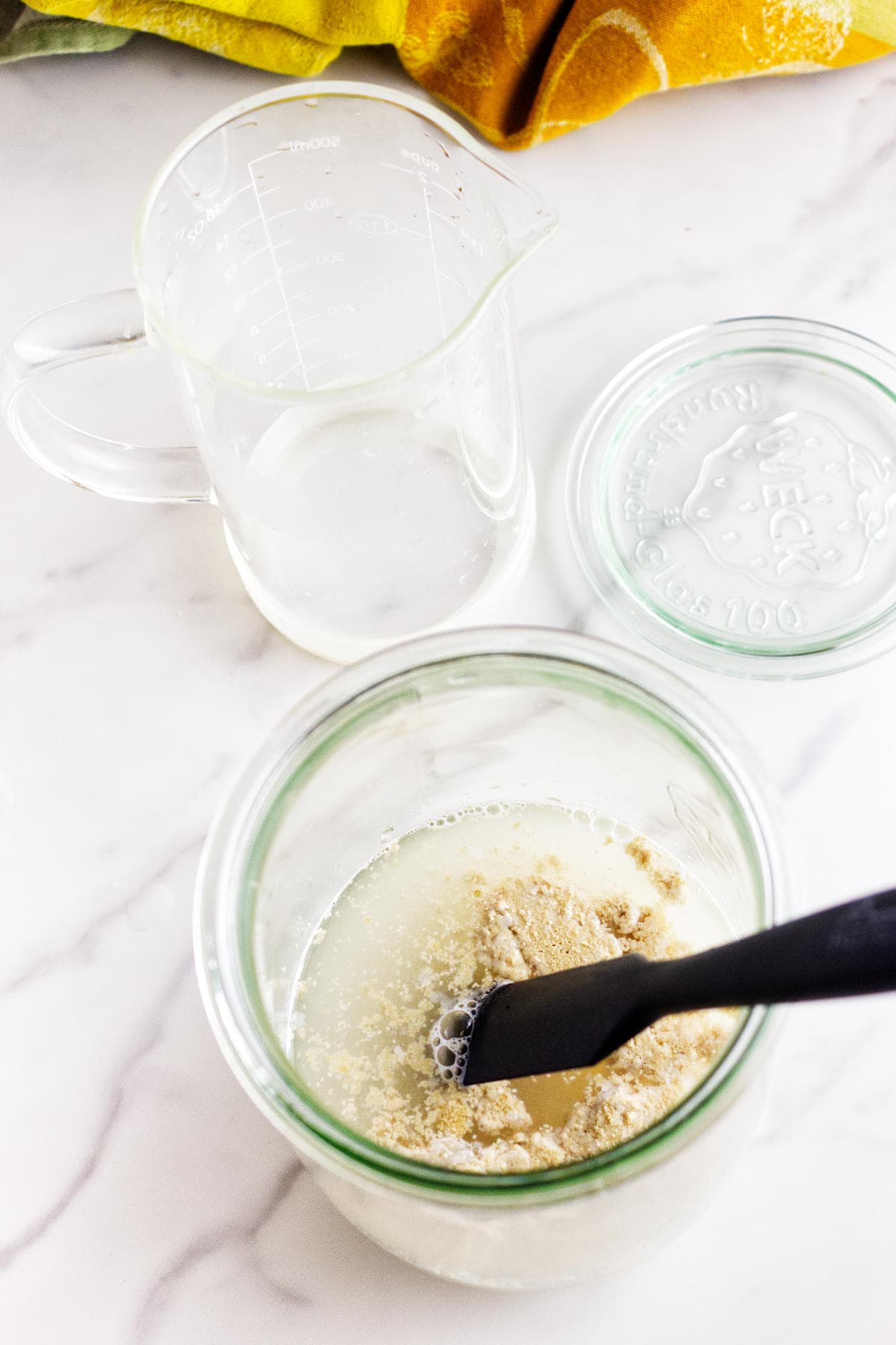 A glass jar with flour, water, and potato flake sourdough bread starter is being mixed with a black spatula on a marble surface. An empty measuring cup and glass lid are nearby, with a yellow towel in the background.